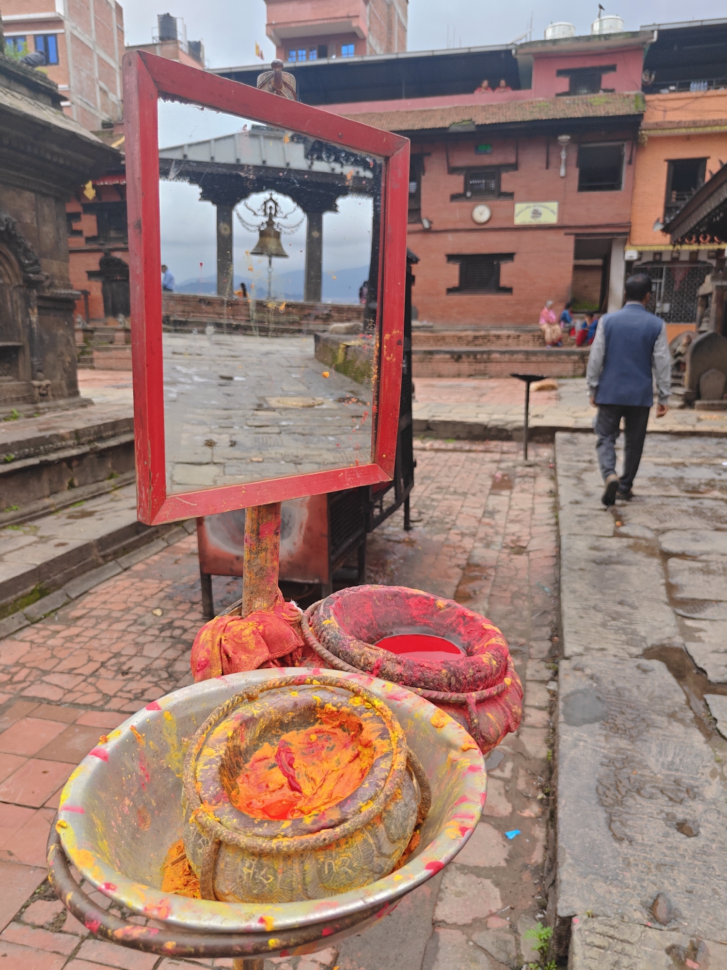 In Kirtipur, Nepal, in front of a brick building with multiple windows and a clock, a red-framed mirror stands upright, reflecting a pagoda-style structure with a large bell hanging from its ceiling. In the foreground, a metal basin holds a red-orange substance, likely a pigment or paste, and is surrounded by red fabric. To the right of the basin, another red object, possibly a bundle of fabric or a ritualistic item, sits on the ground. A man in a blue vest and grey pants walks away from the viewer on a stone-paved path. The ground is wet in places, suggesting recent rain. The overall atmosphere appears to be that of a sacred or historical site, possibly a temple complex, with architectural elements and offerings indicative of religious practice.