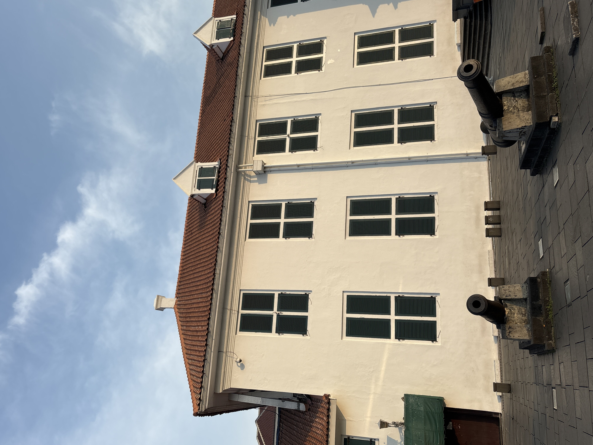 The image depicts the exterior of a white colonial-era building with a red tiled roof. The building has numerous windows with dark green shutters, arranged in neat rows. Two cannons on stone pedestals are positioned in the foreground on a paved area. The sky above is blue with scattered clouds. The scene is likely the Fatahillah Square in Kota Tua, West Jakarta, Indonesia, a historic area known for its Dutch colonial architecture. There are no people visible in the image.