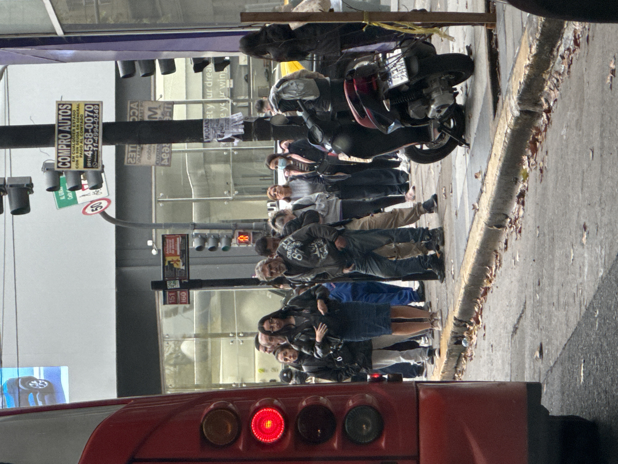 A street scene in Buenos Aires, Argentina, features a red bus with illuminated rear lights in the foreground. A queue of approximately ten people stands on the sidewalk, with a parked motorcycle visible behind them. Signage is present on a pole, including advertisements for "COMPRO AUTOS" (I buy cars) with a phone number and streetcar route numbers. The environment includes a building with large windows, traffic lights, and a general urban street setting.