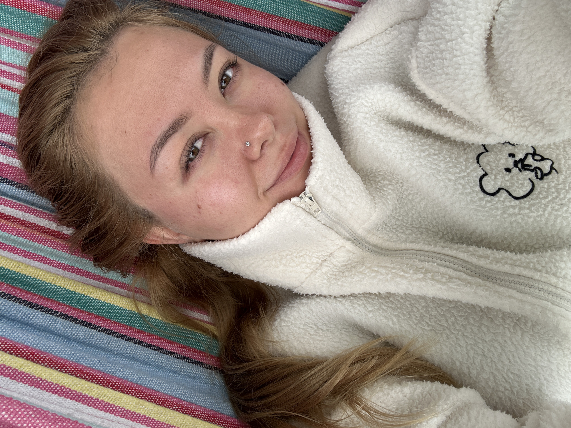 A young woman with light brown hair is lying on a multicolored striped surface, possibly a hammock or blanket. She has fair skin and a nose piercing. She is wearing a white, fleece-like zip-up jacket with a small cartoon-like design on the chest. The background appears to be out of focus. Location: Unknown, Unknown.