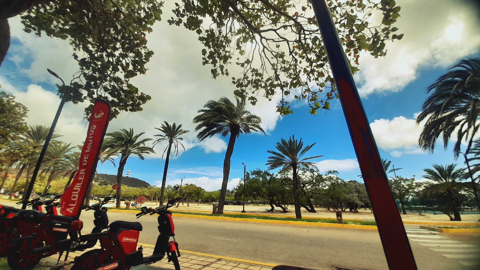 The image shows a street scene in Pedro González, Venezuela, with a row of red electric scooters for rent parked along the side of the road. A red flag with white lettering that reads "ALQUILER DE MOTOS" (Motorcycle Rental) is prominently displayed in the foreground. In the background, several palm trees line the street, and a lighthouse is visible on a distant hill under a blue sky with white clouds. The road appears to be paved, and there is a pedestrian crossing marked with white stripes. The overall impression is one of a tropical or subtropical location, likely a tourist area.