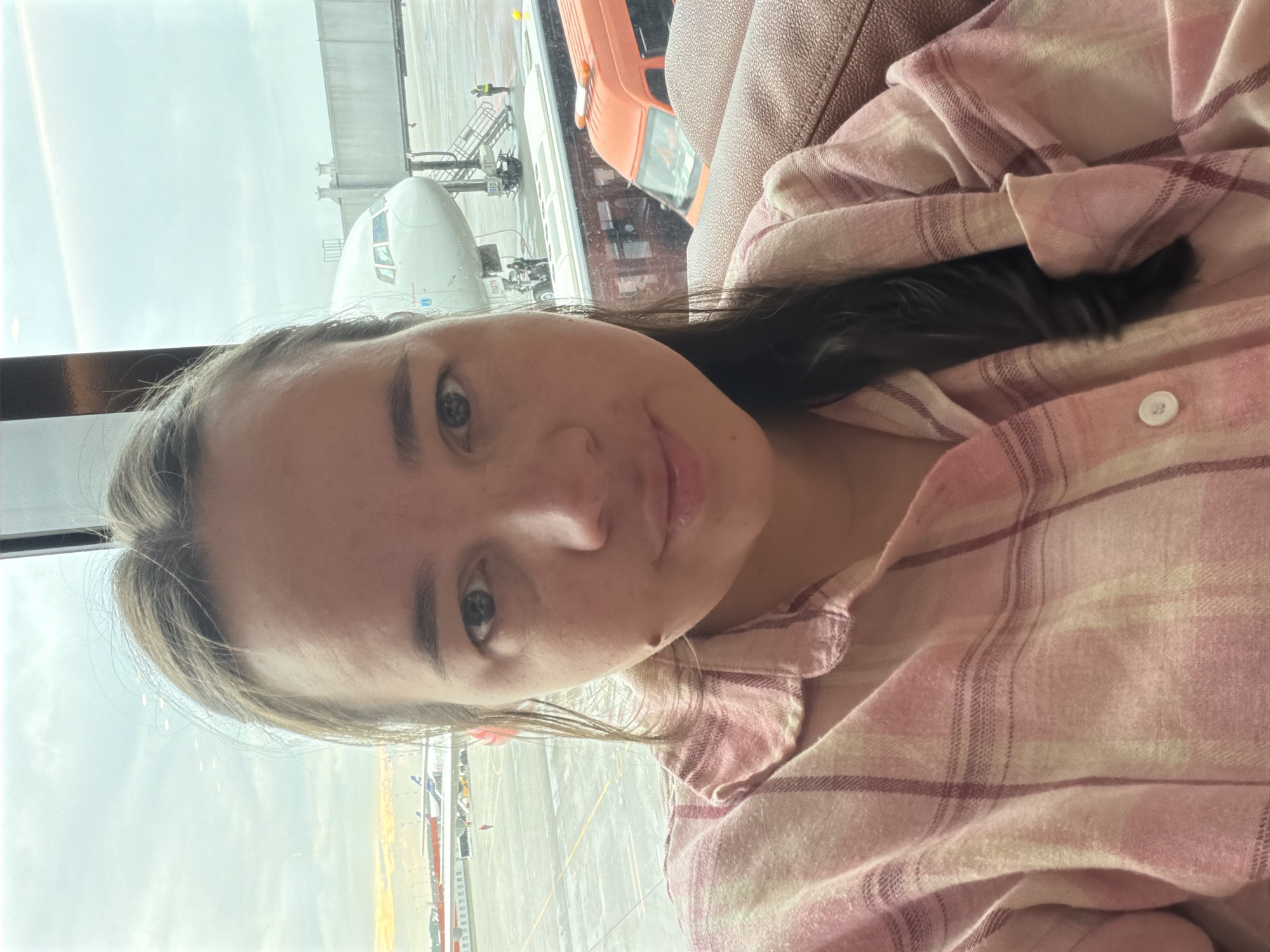A young woman with brown hair and brown eyes is seated indoors, with an airport tarmac visible through a large window. She is wearing a pink plaid shirt. The woman is in the foreground, taking a selfie. Behind her, a large white airplane is parked at a gate, with a jet bridge attached. To the right of the airplane, an orange airport vehicle is visible. The sky outside is overcast, and a distant sunset can be seen on the left side of the image. The location is identified as Khimki, Russia, likely within Sheremetyevo International Airport.