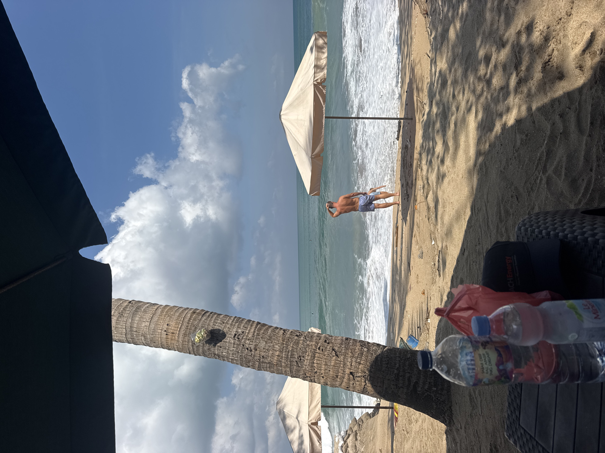 The image depicts a sunny day at a beach in Kuta, Indonesia. In the foreground, on a dark woven table, are two plastic water bottles and a red plastic bag, with a black baseball cap to the right. A large palm tree trunk is visible on the left, with a cluster of green objects near its top. Two beach umbrellas are set up on the sand, one further in the background on the left and another in the middle ground. A man wearing blue swim trunks is standing by the water's edge, facing the ocean and appearing to scratch his head. The ocean is visible with breaking waves and white surf. In the distance, several small figures can be seen in the water, likely surfers. The sky is blue with scattered white clouds. The sand is light brown and appears to have some debris, including plastic, scattered on it. The overall scene conveys a relaxed atmosphere on a tropical beach.