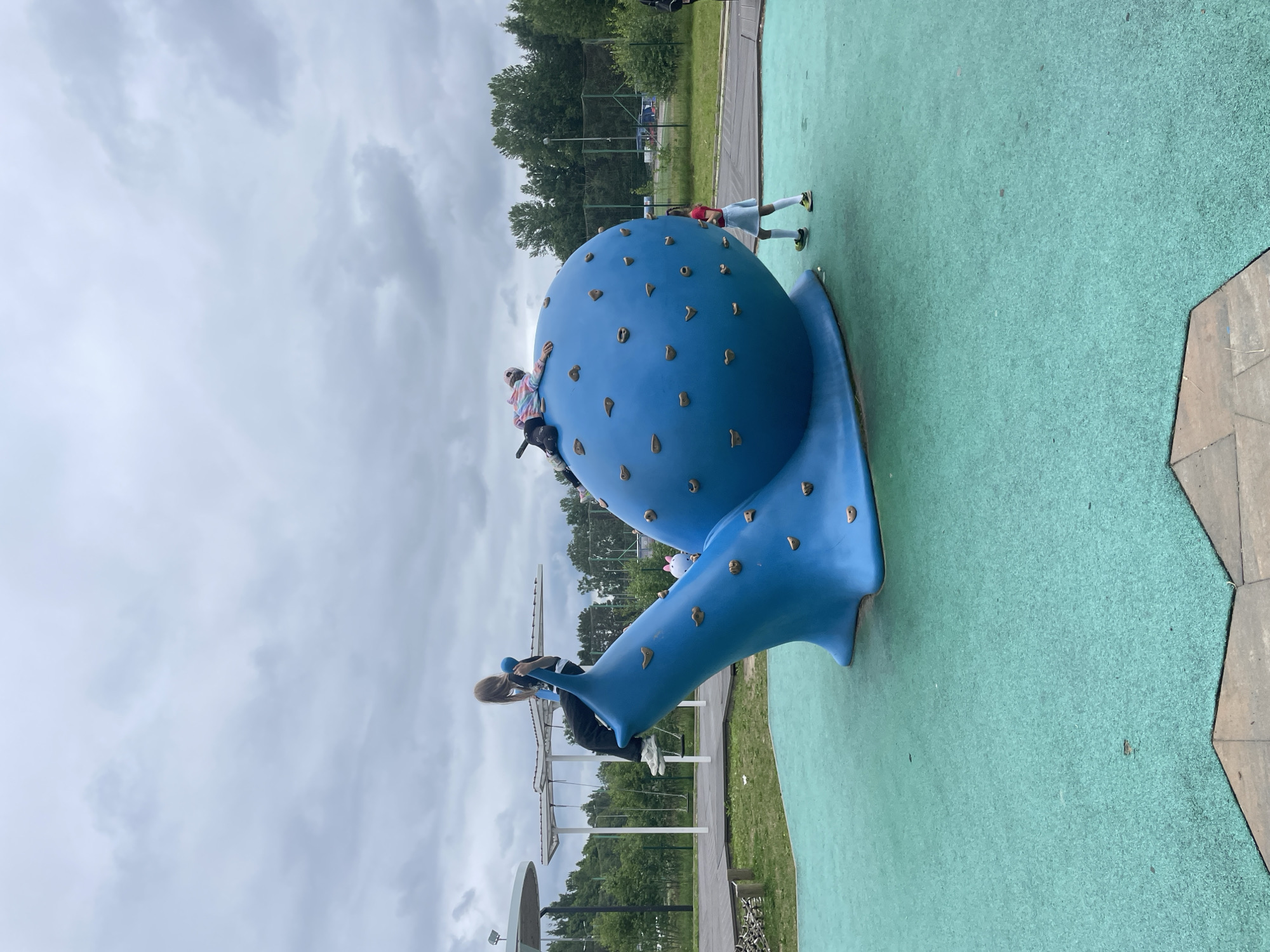 The image shows a playground scene in Sosnoviy Bor, Russia. The main subject is a large, blue, snail-shaped climbing structure with various grips. Several children are actively using the structure. One child in a tie-dye hoodie is climbing on the top part of the snail's shell. Another child, wearing a red shirt and white skirt, is standing on the ground near the snail's body. A third child, with long blonde hair and wearing a black top and dark pants, is on a lower part of the structure, seemingly descending or climbing.
The setting appears to be an outdoor recreational area with a green, soft-surface ground, typical of playgrounds. In the background, there are trees, a sports field with a net, and a cloudy sky. The weather seems overcast, suggesting it might be a cooler day, possibly late spring or summer. The overall mood is playful and active, with the children engaged in climbing and enjoying the playground equipment. There is no visible text in the image. The image is oriented sideways.