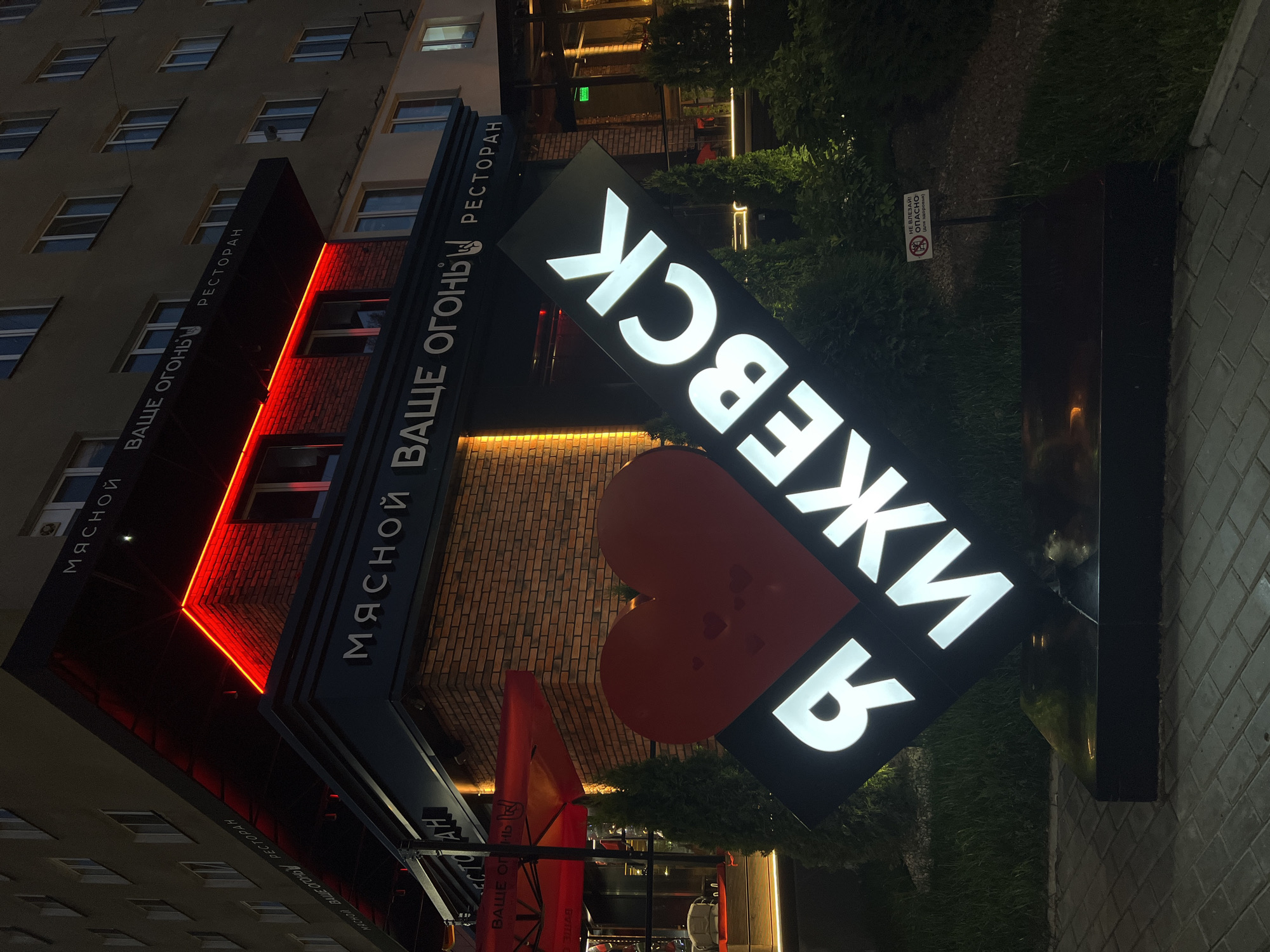 The image shows a night view of a restaurant called "МЯСНОЙ ВАШЕ ОГОНЬ" (Myasnoy Vashe Ogon') in Izhevsk, Russia. The restaurant has a modern facade with red neon lighting and brickwork. In front of the restaurant, a large illuminated sign with a red heart and white text spelling out "Я ИЖЕВСК" (Ya Izhevsk), meaning "I Izhevsk", is prominently displayed. The sign is tilted and rests on a black base. There are several small green trees and bushes around the sign and the restaurant entrance. A small sign with a prohibition symbol and text reading "НЕ ВЛЕЗАЙ! ОПАСНО (дети одёргивают)" (Do not enter! Danger (children pull)) is visible in the background to the right of the main sign. The ground is paved with grey rectangular tiles. The building behind the restaurant appears to be an apartment building with multiple windows. The lighting suggests it is evening or night.