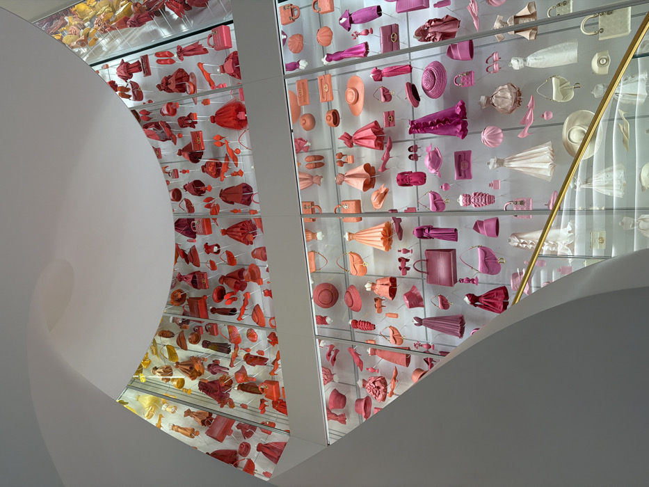 This image captures a multi-story, curved architectural display within the Galerie Dior at 30 Avenue Montaigne, Paris, France. The central focus is a vast exhibition of hundreds of miniature fashion items, meticulously arranged within clear, illuminated display compartments that span the height of the wall. The items are organized in a distinct chromatic gradient: the uppermost sections feature bright reds, oranges, and yellows, transitioning downwards through various shades of pinks, fuchsias, and corals in the middle, to lighter pinks, creams, and whites towards the bottom. The displayed objects include miniature dresses of various styles, an assortment of handbags, different hats, shoes, and other accessories. A smooth, curvilinear white architectural element forms the upper left and top boundary of the frame, indicating the modern interior design. In the lower right, a golden handrail and a partial view of a staircase are visible, suggesting an ascending path alongside the exhibition. No individuals are present in the static scene.