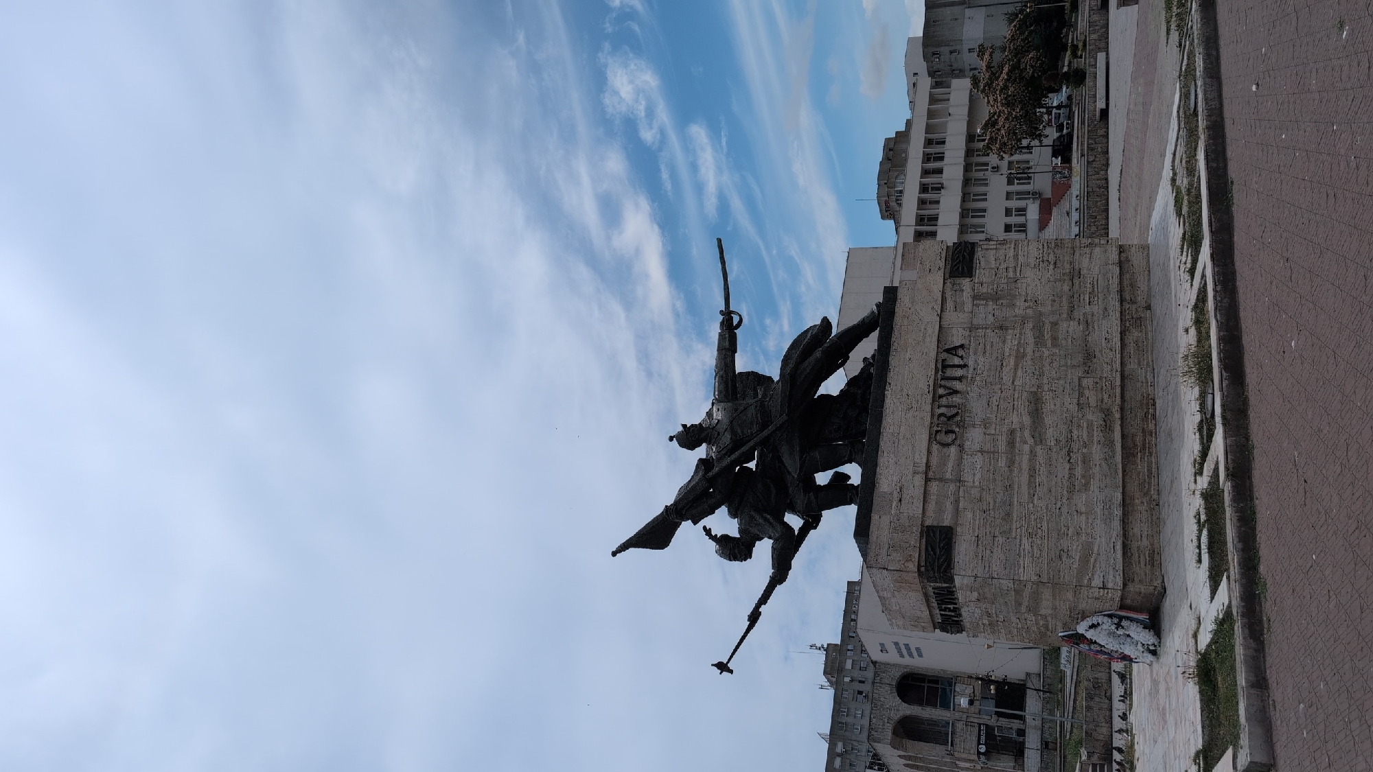The image shows a statue of two soldiers in a city square, likely in Constanța, Romania, given the inscription "GRIVITA" on the monument's base. One soldier, in the foreground and to the right, is depicted wearing a helmet and holding a sword in his right hand, extended forward, and a rifle in his left hand. He is wearing a long coat and appears to be marching or advancing. Behind him and to his left, another soldier, also wearing a helmet, holds a flag in his left hand and what appears to be a rifle or spear in his right hand. The soldiers are mounted on a stone pedestal, which is inscribed with the word "GRIVITA" on one side and "PLEVNIA" on another. A wreath of white flowers is placed at the base of the monument on the left side.
In the background, there are several buildings. To the left, a large, ornate building with arched windows is visible. To the right, a modern, multi-story building with numerous windows and a white facade stands. Further to the right, another building with a darker facade and more windows is seen. Trees, some with pink blossoms, are visible between the buildings on the right. The ground in the foreground is paved with reddish bricks. The sky is partly cloudy with streaks of white clouds against a light blue background. Pigeons are scattered on the ground in front of the monument. The overall scene depicts a historical monument in an urban setting.