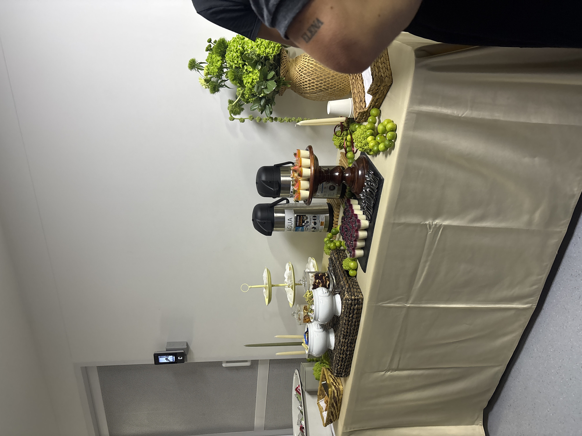 The image depicts a table laden with food and decorations, likely in Bello, Colombia. The table is covered with a light-colored cloth, possibly satin. A woven vase holds green floral arrangements. There are two thermoses, a tiered serving tray with white bowls, and trays of grapes and finger foods. A basket and candles are also present. A person’s arm with a tattoo is visible in the upper right corner of the image. A door with a security camera above it is visible in the lower left. The floor appears to be tiled.