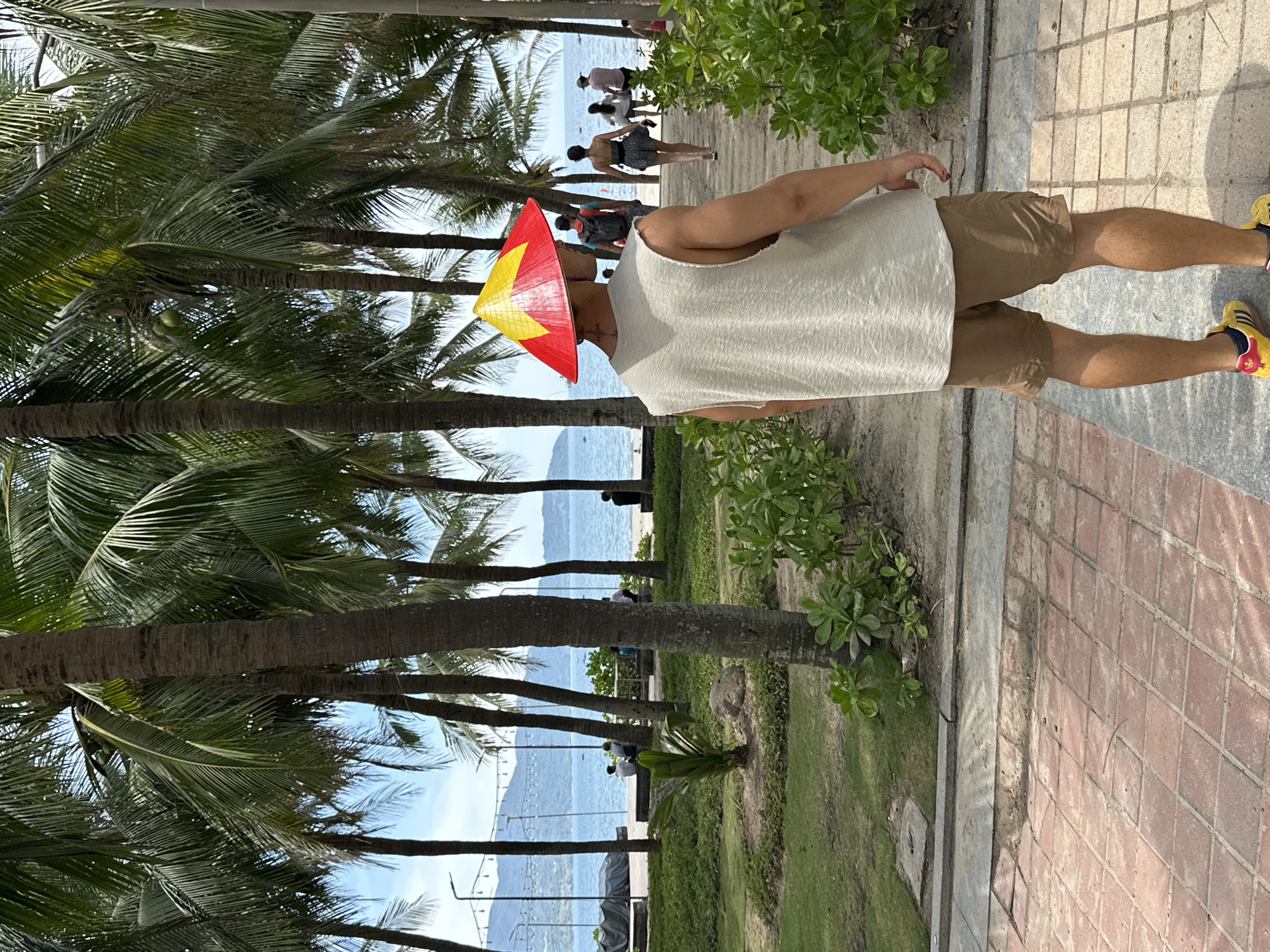 The image shows a person wearing a conical hat with the Vietnamese flag design, standing on a paved walkway lined with palm trees. The person is wearing a light-colored tank top and khaki shorts, with yellow and black athletic shoes. The background features a beach with people walking in the distance and the ocean. The setting appears to be a tropical or subtropical location, with lush greenery and palm trees. The time of day seems to be daytime, with sunlight casting shadows. The overall mood is relaxed and casual, suggesting a vacation or leisure activity. No text is visible in the image. The location context is Đà Nẵng, Vietnam.