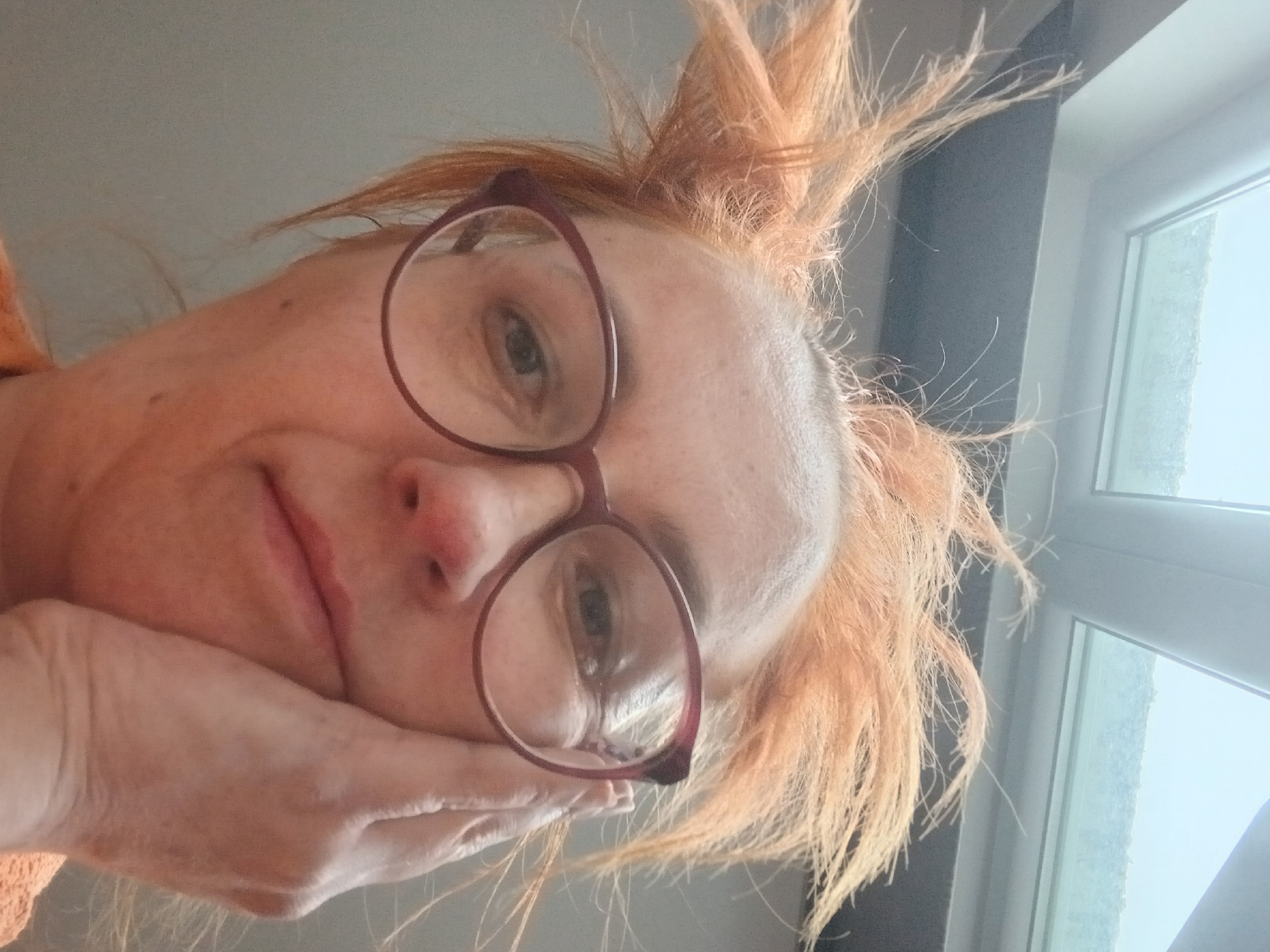 A close-up, selfie-style portrait shows a woman with reddish-blonde hair, styled upwards, wearing round, burgundy glasses. Her face is supported by her left hand. The background includes a window frame. There is no explicit information indicating that the image was taken in Riga, Latvia.