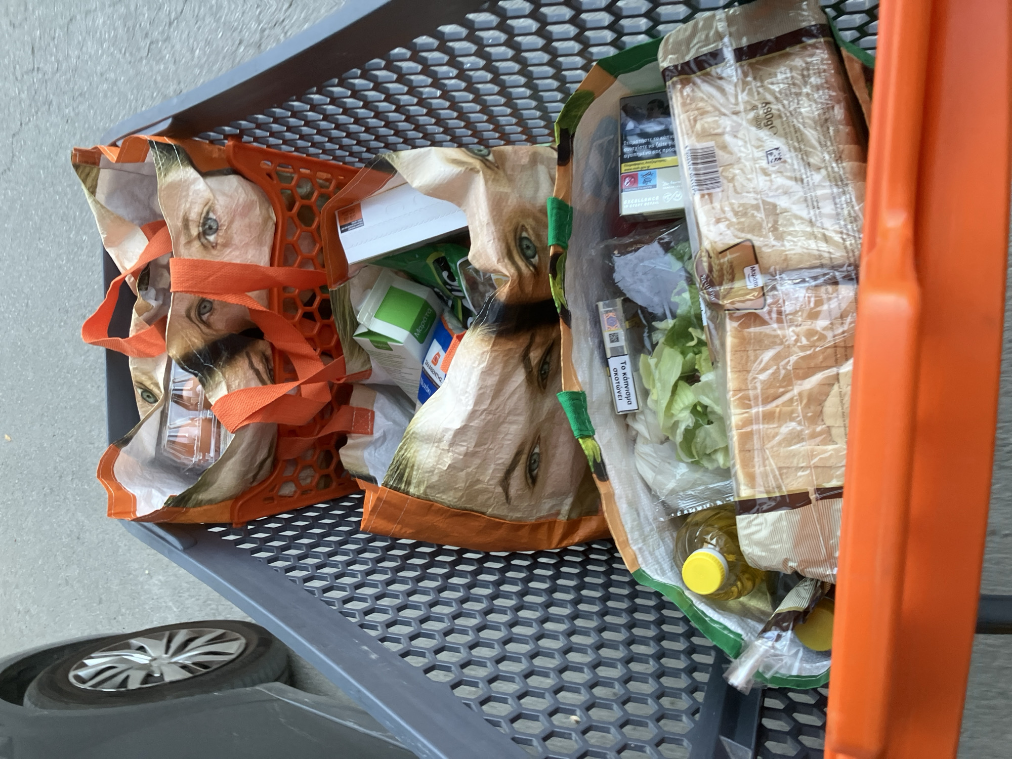 A high-angle, full shot captures a shopping cart filled with groceries. The cart is a dark gray plastic with a honeycomb-patterned interior. The groceries are contained within three reusable shopping bags, two of which are orange and feature a close-up print of a person's eyes and forehead.
The top bag contains a clear plastic container with two brown eggs. Below this, the second bag is visible, also with the eye print. Inside this bag, various items are packed, including a green-and-white box, a small white box, and some other indistinct items.
The bottom bag is partially obscured by the orange handle of the shopping cart. It contains a bottle of yellow liquid, likely olive oil with a yellow cap, and a head of lettuce. Adjacent to the lettuce is a plastic-wrapped loaf of sliced white bread. Other items in this bag are less clear but appear to include a packet of cigarettes.
To the left of the cart, a portion of a dark gray car, including its tire and wheel rim, is visible. The ground appears to be paved, suggesting an outdoor location such as a parking lot or sidewalk. The lighting indicates it is daytime.