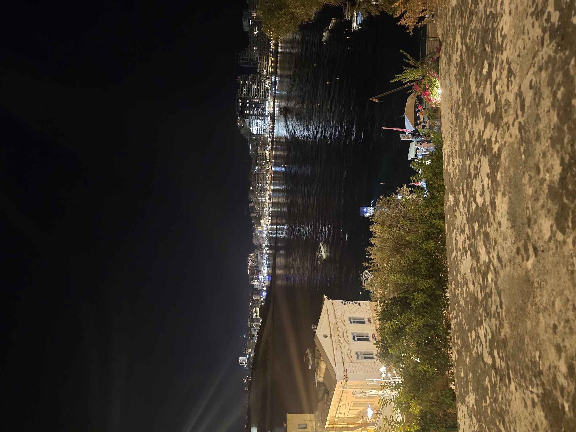 This is a nighttime, high-angle, long shot of a waterfront in Valletta, Malta. The foreground consists of a textured stone surface with shadows from foliage. Beyond this, a lush green hedge lines the edge of the view. In the midground, a cream-colored building with classical architectural details, possibly a restaurant or establishment, is illuminated by lights. The building has multiple windows and an arched entryway. A body of dark water, reflecting the city lights, stretches across the frame. Several small boats are visible on the water, some anchored and one in motion. The background features a cityscape with numerous buildings of varying heights, all lit up, creating a bright and lively urban panorama against the dark night sky. The city lights are reflected on the water's surface, creating shimmering streaks. There is a slight lens flare in the upper left corner of the image.