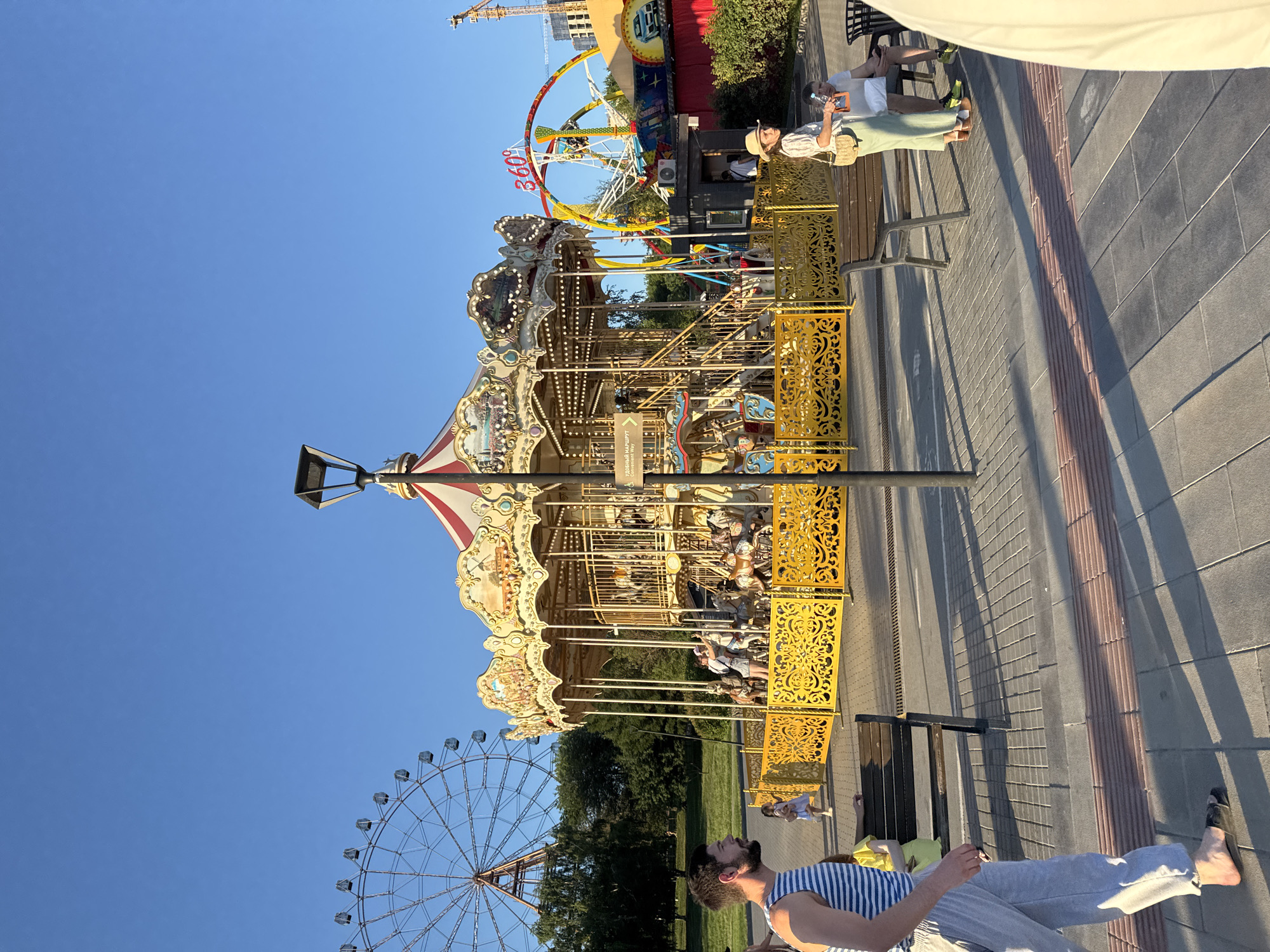 The image captures a scene at an amusement park on a sunny day, likely in Novosibirsk, Russia. The main focus is a vibrant carousel with a red and white striped canopy, adorned with intricate golden railings. Several people are visible enjoying the attractions. In the background, a large Ferris wheel stands tall against a clear blue sky, and another thrilling ride with a circular structure and red lettering that says "360" is also present.
On a nearby bench, two individuals are seated. One person, wearing a straw hat and a light-colored dress, is holding a phone. Another person, wearing a white shirt, is seated next to them. In the foreground, a man with a beard wearing a striped tank top and grey pants is looking upwards. The ground is paved with gray tiles, and the scene is bathed in warm sunlight, casting long shadows, indicating it is likely late afternoon or early evening. The overall atmosphere appears relaxed and cheerful, with families and individuals enjoying a day out. The presence of the carousel, Ferris wheel, and other rides firmly places the scene within an amusement park setting.