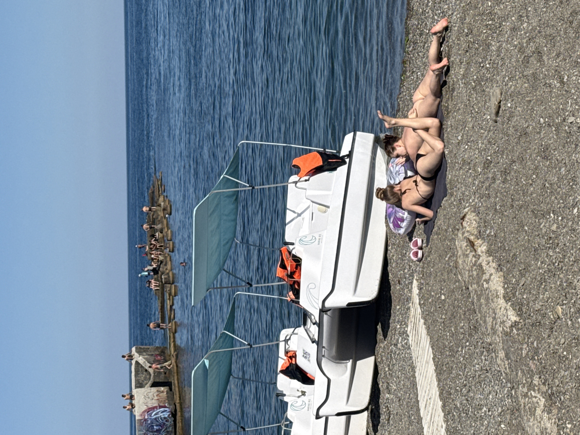 The image depicts a sunny day at a coastal location in Alushta, Ukraine. The scene is divided between the sea and a pebble beach. In the foreground, two white pedal boats with blue awnings are parked on the beach. One of the boats has an orange life vest on it. Further down the beach, three young women are sunbathing on a towel. One woman is lying on her back with her legs up, while the other two are lying face down. They are all wearing bikinis.
In the background, there is a man-made structure extending into the sea, possibly a pier or breakwater, with many people sunbathing and swimming. The water is calm and a deep blue. The sky is clear and blue, suggesting fair weather. The time of day appears to be midday or early afternoon due to the strong sunlight and shadows. The overall mood is one of relaxation and leisure. There is graffiti on the concrete structure in the background. There is no visible text on the boats or other objects in the image.