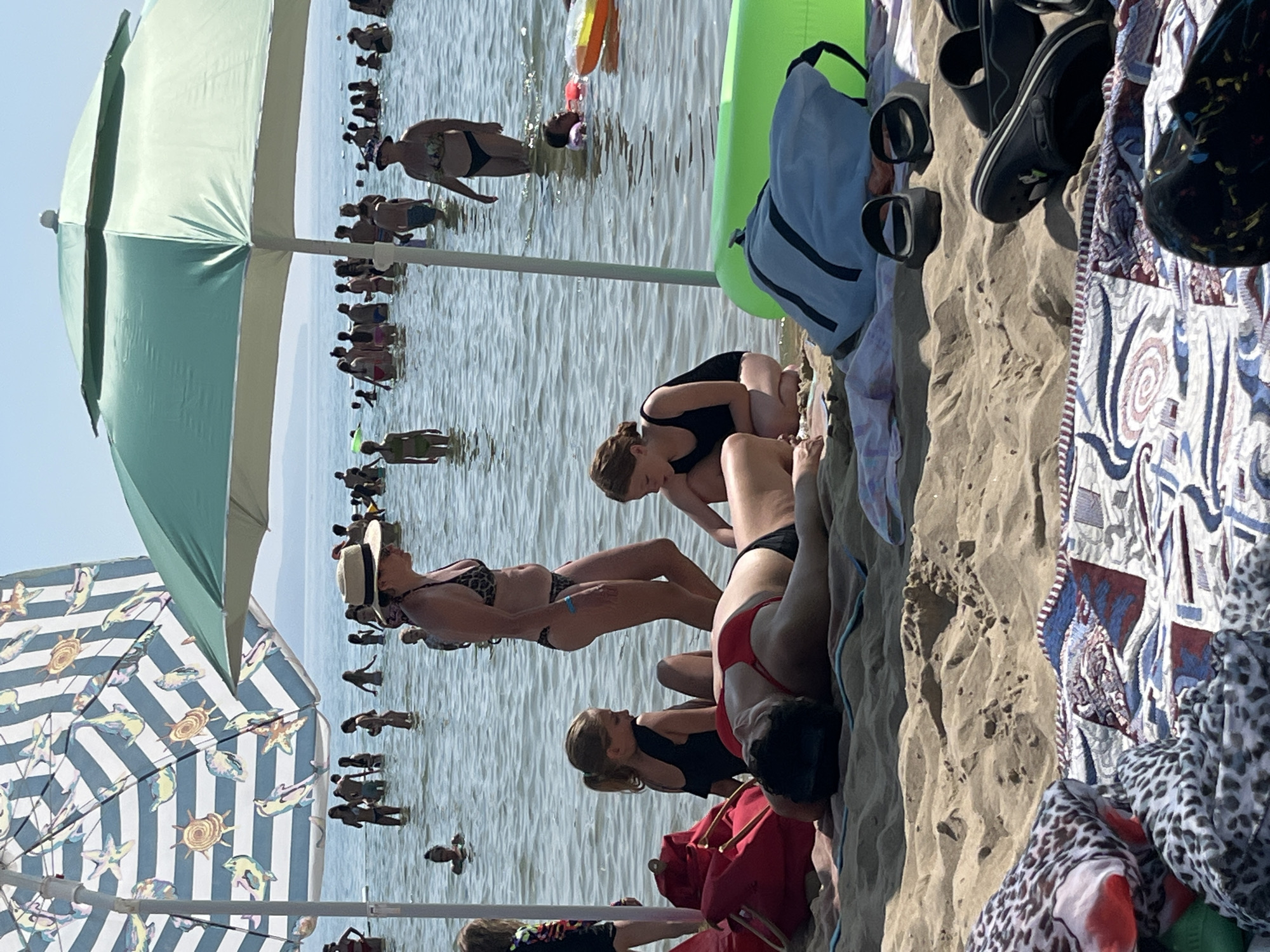 This image depicts a lively beach scene, likely in Nesebar, Bulgaria, during what appears to be a sunny day. The primary subjects are numerous people enjoying the water and the beach. Several individuals are wading in the shallow sea, while others are relaxing on the sand.
In the foreground, the edge of a beach umbrella with a distinctive blue and white striped pattern featuring sea creatures like starfish and shells is visible. Next to it, another umbrella, partially open, is mint green with white trim. On the sand, several beachgoers are seated or lying down. One woman in the foreground is wearing a red bikini top and black bottoms, lying on her side, with her face turned away. Another woman in a black swimsuit is seated next to her. Further back, a woman in a black bikini is lying on the sand, and another woman in a patterned bikini is also on the sand.
In the water, a group of people are standing and walking. Some are seen from the waist up, engaged in various activities. A bright green inflatable is also present in the water. The water itself is a calm, blue-green, reflecting the sunlight. The sand appears light brown and textured.
The overall mood is one of leisure and summer enjoyment, with people taking advantage of the beach and sea. The time of day seems to be midday, given the brightness and shadows. The presence of beach umbrellas and the attire of the people strongly suggest a recreational setting. No visible text is present in the image.