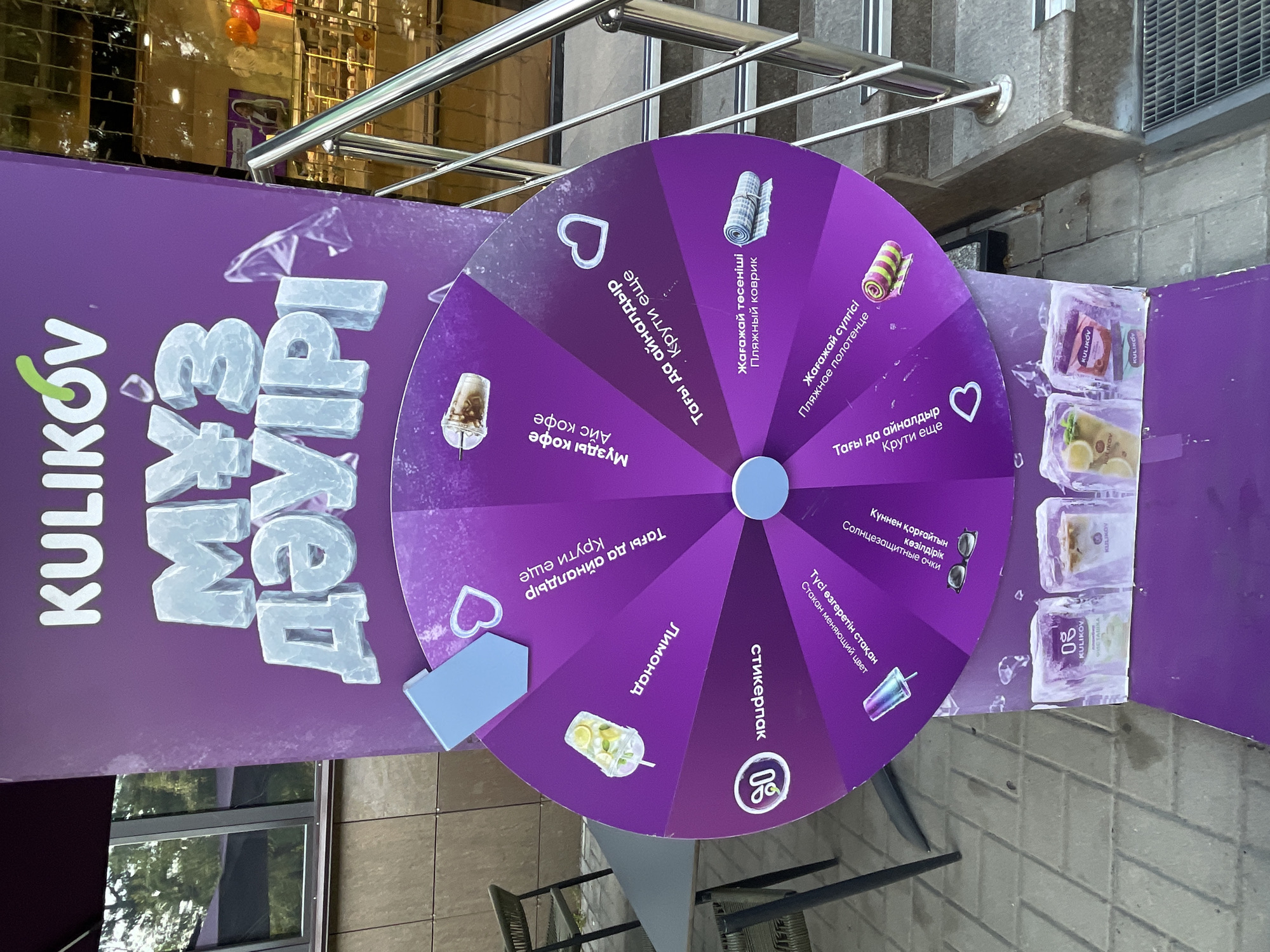 A purple promotional stand for "KULIKOV" ice cream is set up outdoors on a sidewalk in Almaty, Kazakhstan. The stand features a large spinning wheel with various prizes depicted and text in Cyrillic and Russian. The prizes include a lemonade, an iced coffee, a beach towel, sunglasses, and a cup that changes color. The wheel has an arrow indicating where it should stop. In front of the stand, four ice blocks containing different ice cream products are displayed. The background shows a building with windows and a staircase with a metal railing. A table and chair are visible to the left of the stand. The ground is paved with grey tiles. No people are visible in the image.