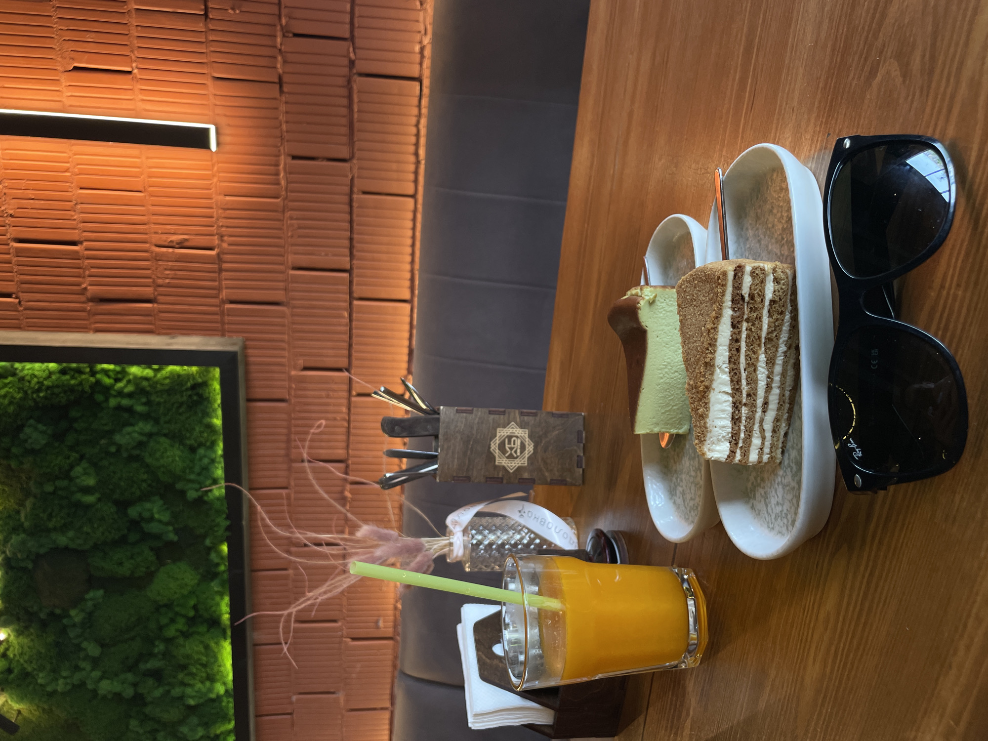 The image captures a scene on a wooden table inside a building in Kazan, Russia. On the table, there is a glass of orange juice with a straw, a tissue holder containing napkins, a small vase with dried flowers, a wooden box containing cutlery, and a plate with two slices of cake. Next to the plate, there is a pair of black sunglasses. In the background, there is a textured orange brick wall with a long rectangular light fixture and a green moss wall art with a black frame. A grey, padded bench extends from the middle to the top of the frame.