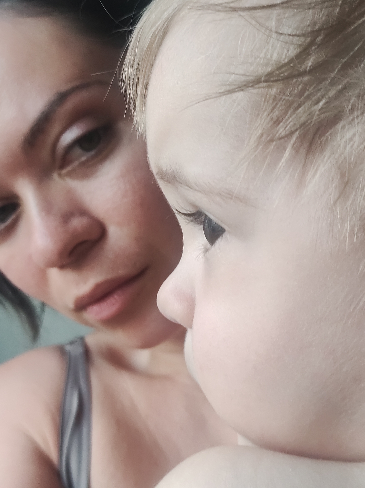 A close-up shot shows a woman and a young child. The woman, with dark hair pulled back and subtle makeup, is gazing downwards with a slightly pensive expression. The child, with light blonde hair, is positioned in the foreground, facing to the right, with the woman behind them. The child's eye is clearly visible, appearing dark against their fair skin. The background is indistinct but suggests a soft, natural light. No indication of the city of Chaikovsky, Russia, can be discerned from the image.