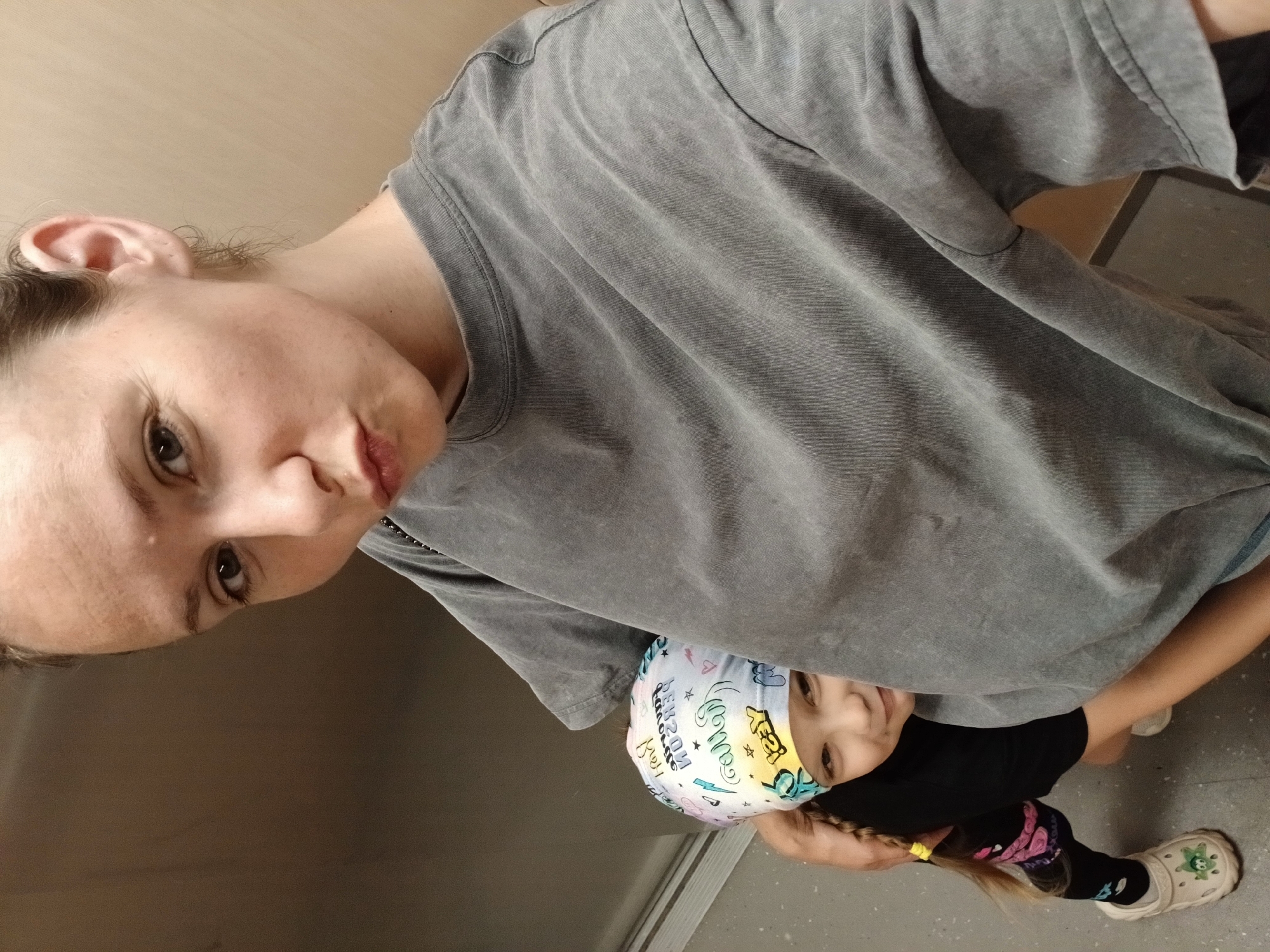 A selfie shows a woman and a child. The woman is in the foreground, with her face turned towards the camera. She has her lips pursed and her eyes are looking to the side. She is wearing a grey t-shirt. Partially visible behind her is a child, who is wearing a bandana with colorful text and drawings. The child's face is visible, and they appear to be smiling. The background is a neutral, indoor environment, possibly a hallway or elevator. The image appears to have been taken in Yekaterinburg, Russia.