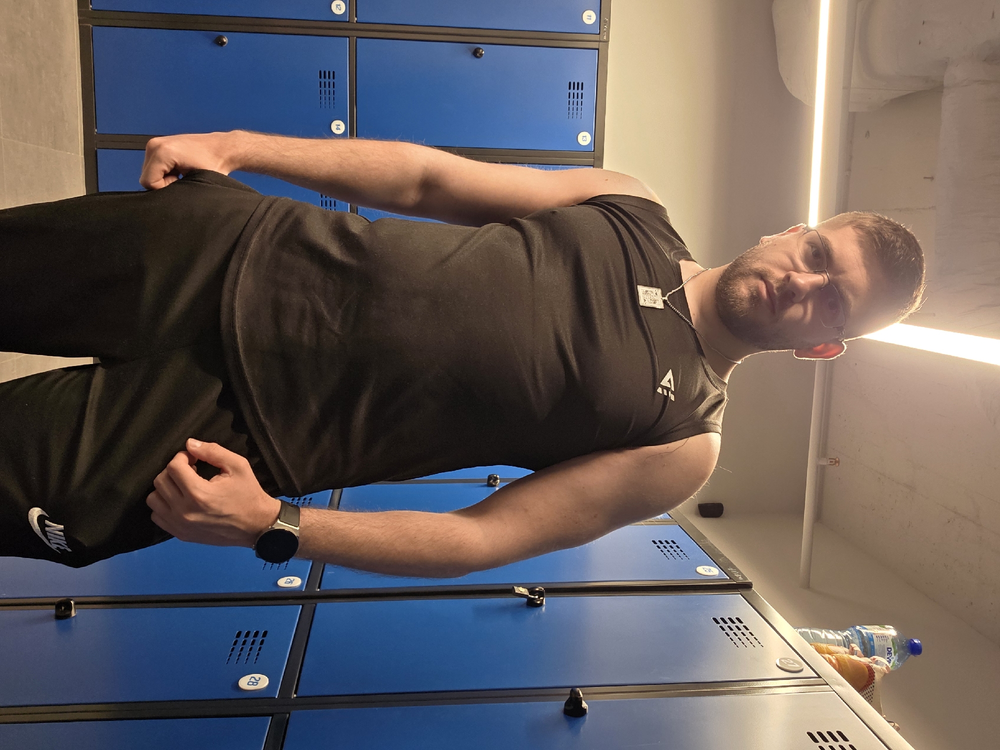 A man stands in what appears to be a locker room, evidenced by rows of blue lockers with black handles and numbering visible. He is dressed in a black sleeveless shirt with a logo and black Nike sweatpants. He wears a wristwatch on his left wrist and a necklace. The man is positioned sideways to the camera with his right hand near his hip and his left hand slightly extended. A water bottle is sitting on top of the lockers near the man.
Without additional information, location within Plovdiv, Bulgaria, cannot be determined.