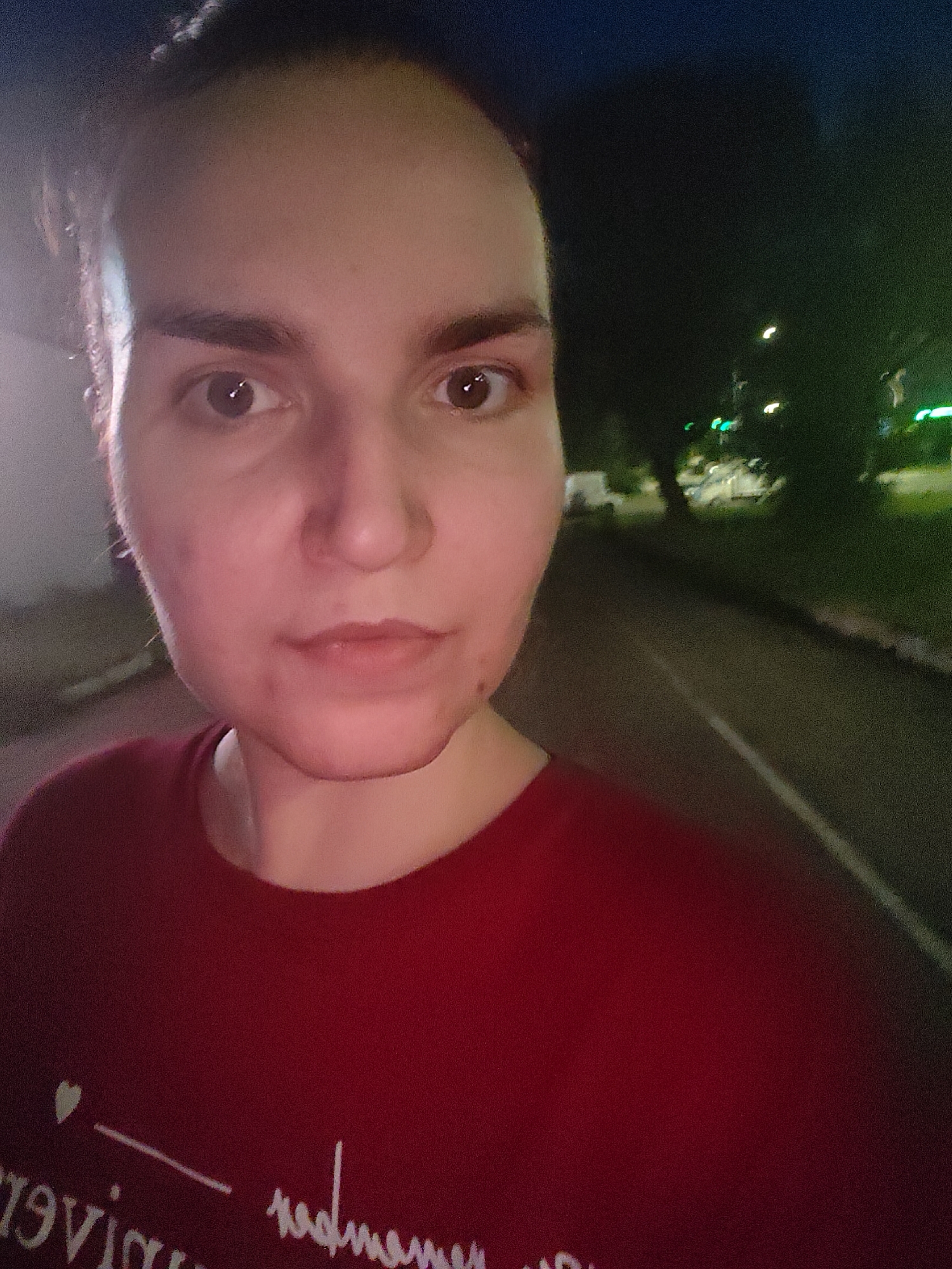 A selfie of a person taken at night. The person is in the foreground, facing the camera with a neutral expression. They have light skin, brown eyes, and dark hair pulled back. They are wearing a dark red t-shirt with some white text and a small heart graphic. The background is blurred, showing a road or path with some greenery and streetlights, indicating an outdoor setting. The lighting suggests it is nighttime.