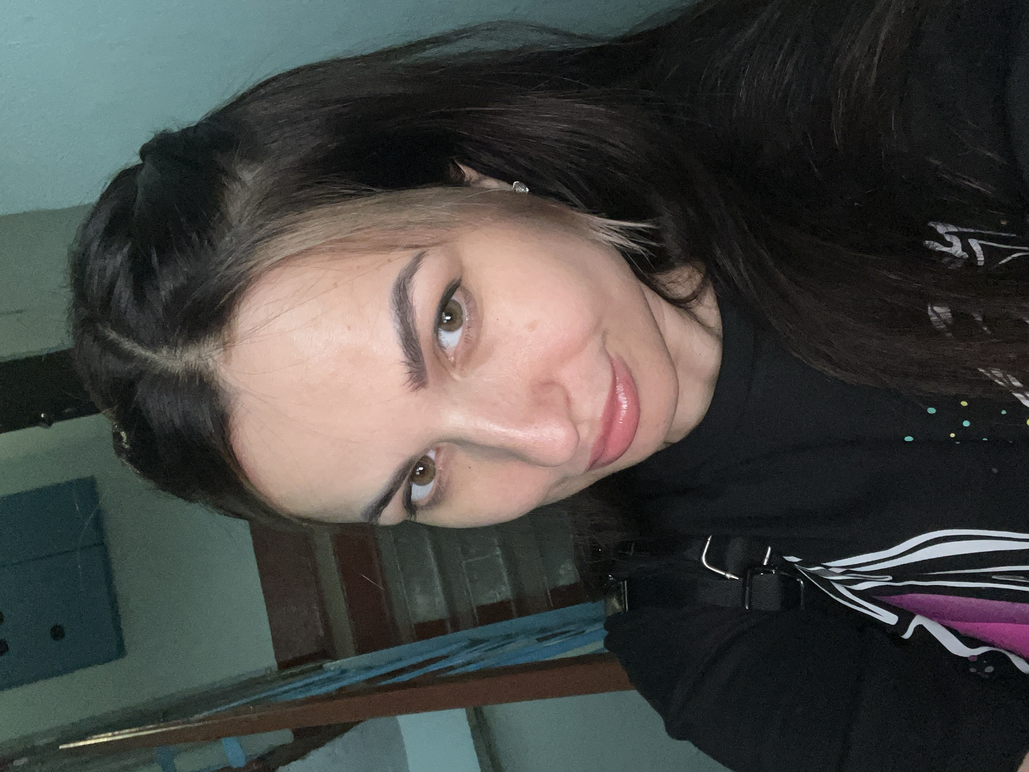 A close-up portrait shows a woman with long, dark hair that has lighter, streaked sections framing her face. Her brown eyes look gently towards the viewer, and she has a soft, closed-mouth smile with light pink, glossy lips. Her eyebrows are well-defined. She is wearing a black long-sleeved top or sweatshirt, which features a design of black and white stripes transitioning into a pink and purple gradient on the right side of her chest. A metallic buckle and a black strap are visible near her left shoulder, suggesting part of a bag or accessory. A small, subtle silver hair clip is seen in her hair near her temple. The background is out of focus, revealing muted blue-green walls and darker brown/reddish architectural elements, possibly indicating an indoor setting such as a hallway or stairwell. The lighting appears to be dim. No text is visible.