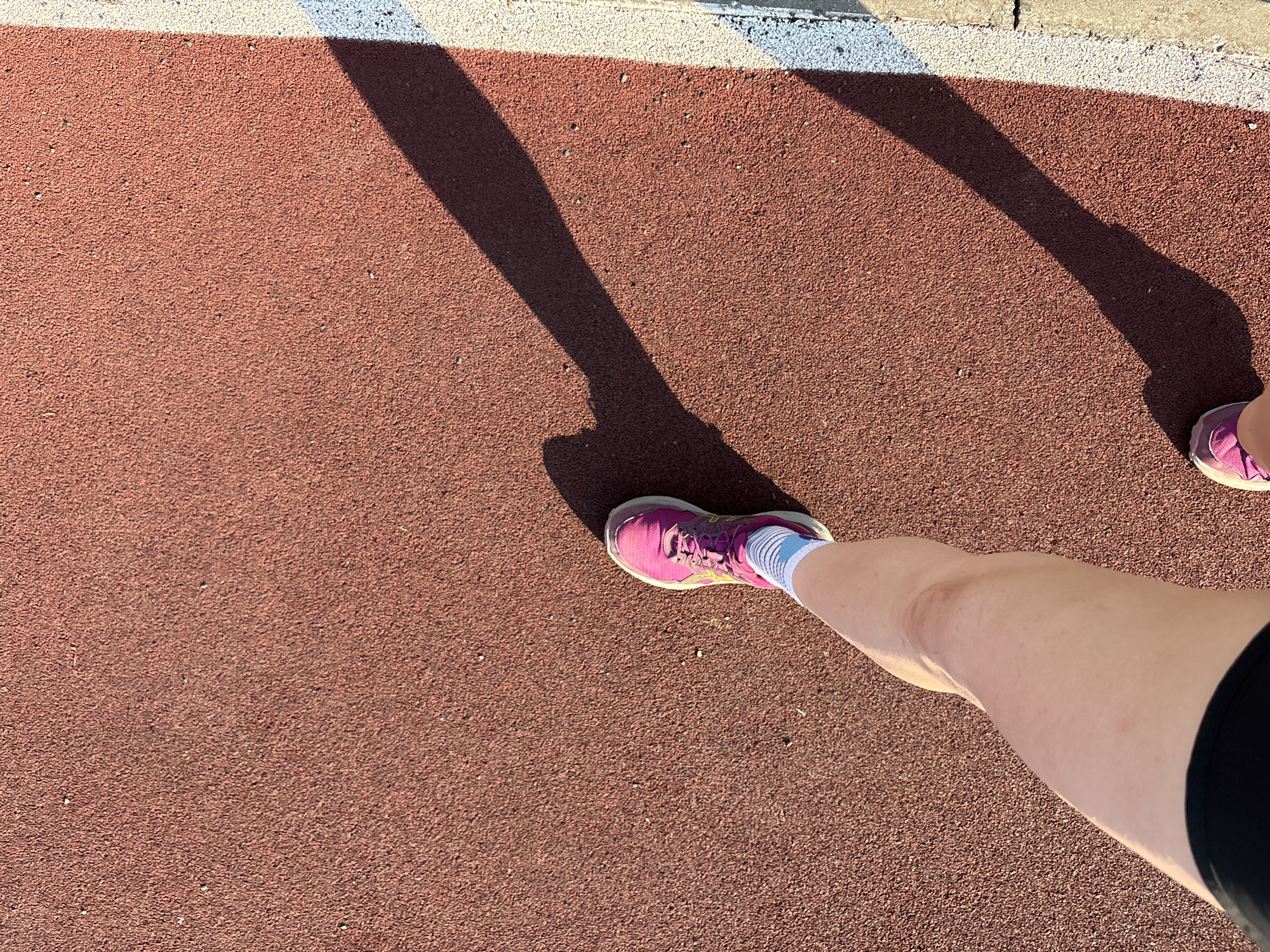 A high-angle shot shows a portion of a red running track, likely outdoors, with the legs of an individual standing on it. The person is wearing pink running shoes, white socks, and dark-colored athletic shorts. Their legs and the shadow cast by their body are visible on the track. The running track has a textured, rubbery surface. A strip of white and gray marking the edge of the track can be seen at the top of the frame. There is no information in the image to confirm its location in the city of Perm, Russia.