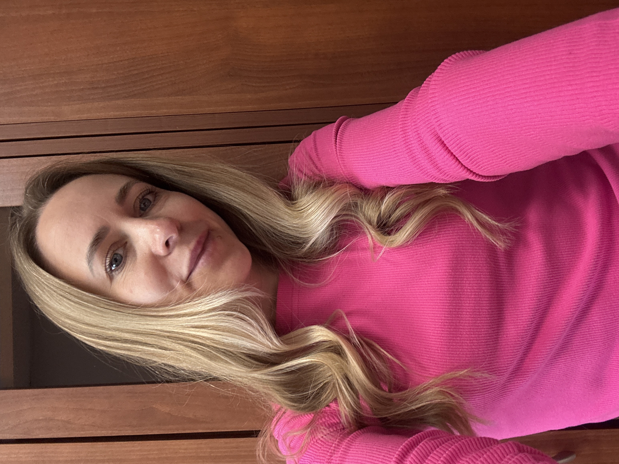 A woman with long blonde hair and blue eyes is wearing a bright pink long-sleeved shirt. She is positioned indoors, near a wooden structure, possibly a cabinet or wall. The background is a light-colored wood, seemingly indoors. There is no indication of the location being Chaikovsky, Russia.