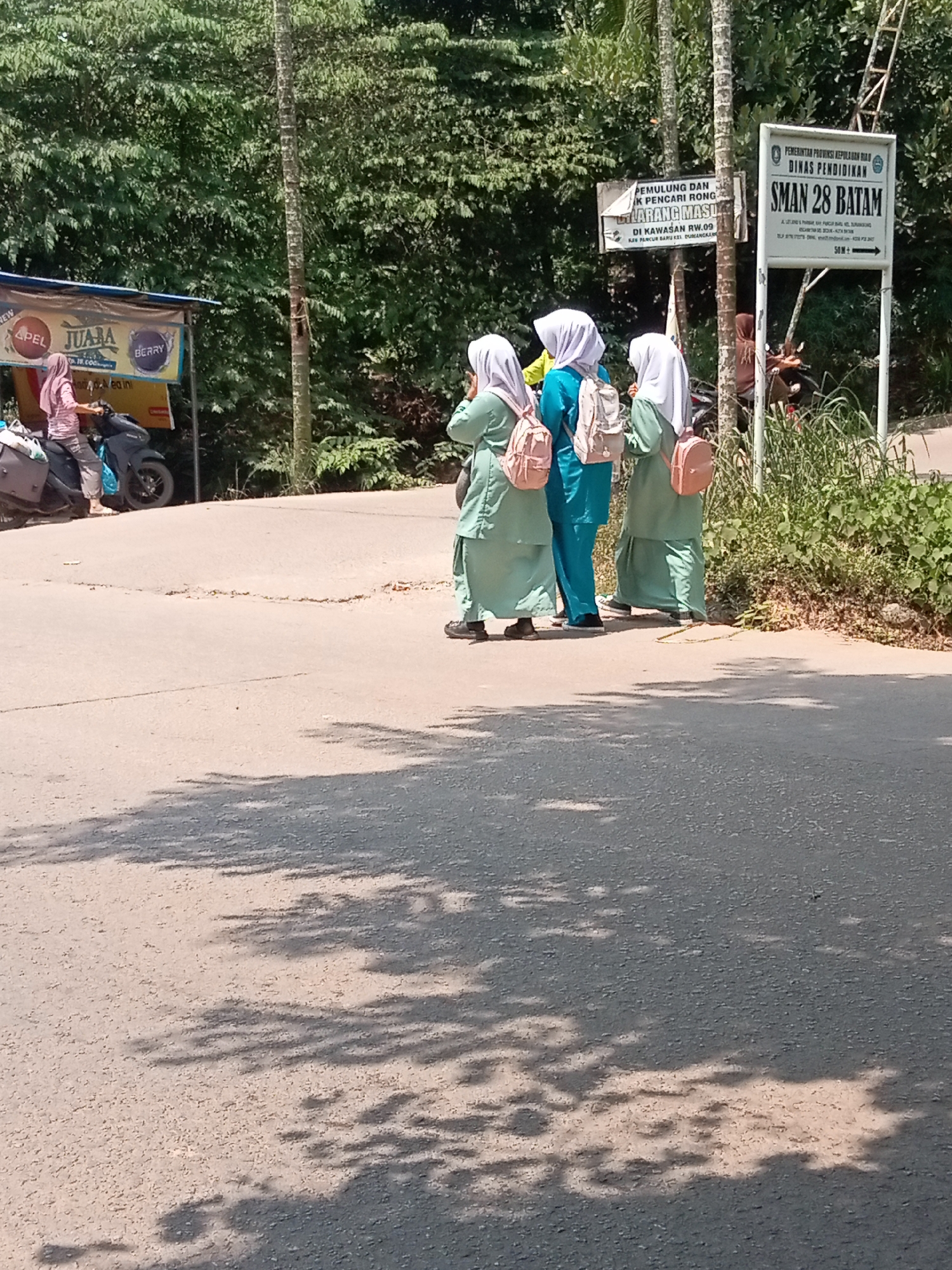 The image depicts a roadside scene in Batam, Indonesia, likely near SMAN 28 Batam. Three young individuals wearing light green uniforms and hijabs are standing on the side of an asphalt road, facing away from the camera. They are carrying backpacks. To the left, a person in a pink hijab is seated on a dark-colored motorcycle, which is parked near a vendor's stall displaying products like "APEL JUARA" and "BERRY." The stall has a blue awning. In the background, lush green trees and foliage line the road. A white sign with text, including "PEMERINTAH PROVINSI KEPULAUAN RIAU DINAS PENDIDIKAN" and "SMAN 28 BATAM," is visible to the right, indicating the location of the school. Another smaller sign with "50 M+" suggests the school is a short distance away. The road surface shows signs of wear and tear, with cracks and some rough patches. Sunlight casts shadows of trees onto the road, suggesting it is a bright day.