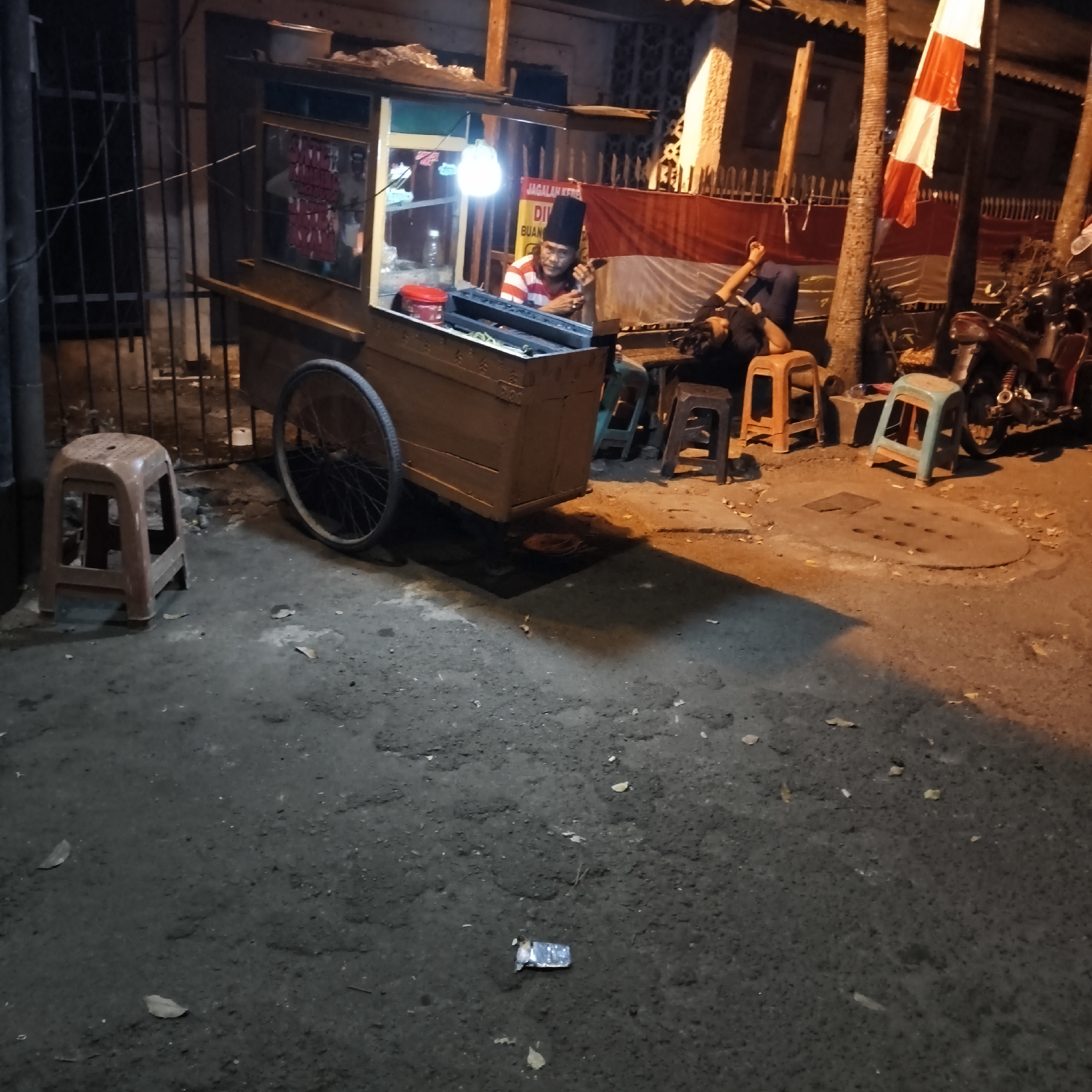 The image depicts a street scene at night in South Jakarta, Indonesia, likely in a neighborhood with food stalls. In the foreground, a dirt and asphalt ground is visible with scattered leaves and a piece of trash. To the left, a small brown plastic stool sits on the ground. In the midground, a food cart is positioned, featuring a wooden structure with a transparent display window. Inside the cart, a person wearing a red and white striped shirt and a tall black cap is seated, appearing to be the vendor. They are holding a phone to their ear and looking towards the viewer. Behind the vendor, a red and white Indonesian flag is displayed. To the right of the food cart, another person is lying down on several plastic stools, with their arm extended. There are several more plastic stools of various colors (orange and light blue) arranged near the food cart and the person lying down. In the background, a motorcycle is parked and partially visible. The environment is dimly lit, suggesting it is evening or night, with artificial lighting from the food cart and ambient streetlights casting shadows. The overall scene suggests a casual, perhaps late-night, setting at a local eatery.
