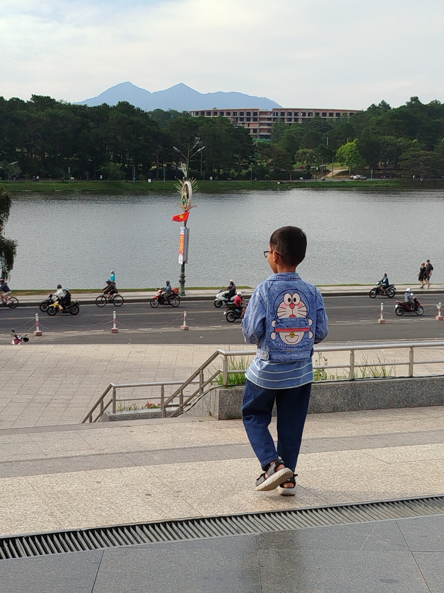 A boy stands with his back to the viewer, wearing a denim jacket with a Doraemon graphic, blue striped shirt, and jeans, gazing at a lake in Da Lat, Vietnam. A road lines the lake with motorbikes and bicycles traveling along it. Beyond the road, a building is partially obscured by trees, and mountains rise in the distance under a cloudy sky. The boy stands on a tiled terrace with metal railings. A Vietnamese flag is visible on a pole near the road.