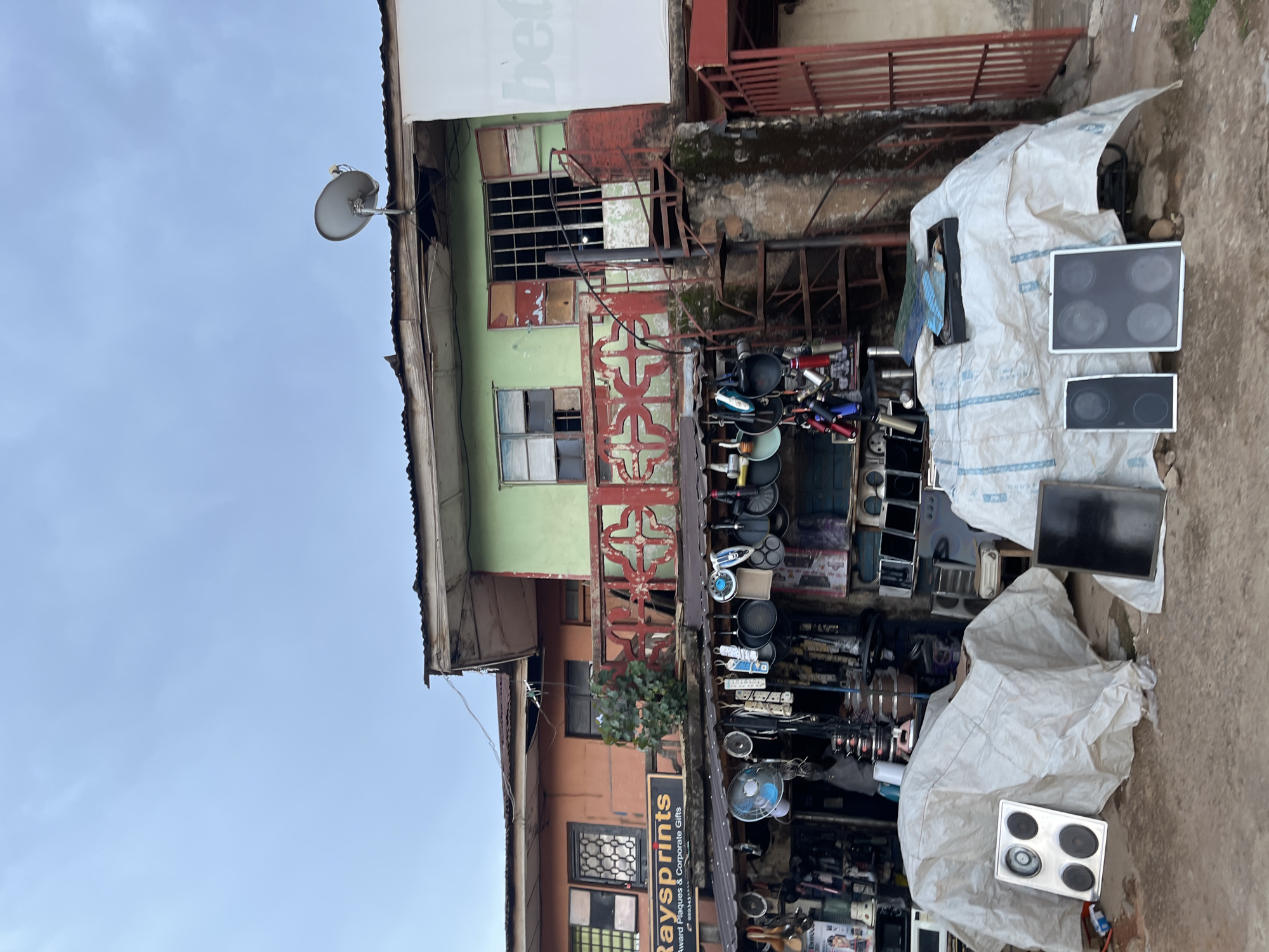 The image depicts an outdoor market scene in Jos, Nigeria, characterized by a cluttered and eclectic display of goods. The focal point is a shopfront with a faded orange and green facade, exhibiting a mix of household appliances, kitchenware, and other assorted items. A prominent sign above the shop reads "Raysprints Award Plaques & Corporate Gifts."
In the foreground, several electrical appliances, including stovetops and what appear to be blenders or food processors, are covered with white tarpaulins, suggesting they are either for sale or in storage. Other items, such as fans, pots, pans, and extension cords, are hung from hooks and displayed on shelves along the shop's exterior. A spiral metal staircase ascends the side of the building, leading to an upper level with windows and a satellite dish on the roof.
The environment appears to be a street or pathway, with rough ground and debris visible. The overall impression is one of a busy, informal marketplace with a diverse range of merchandise. The sky is overcast, indicating a cloudy or potentially rainy day. No people are visible in the frame.