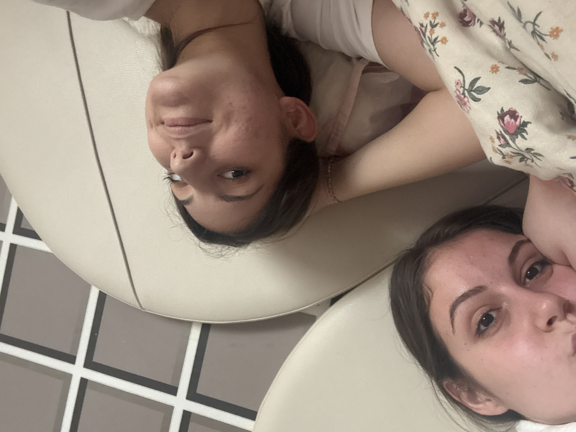 This indoor photograph features two young women relaxing side-by-side, looking towards the camera. The image is presented such that their heads are towards the top of the frame, and their bodies extend downwards, implying they are lying on their backs.
The woman on the left has dark hair and fair skin, with some minor blemishes visible on her cheek. She wears a white top and a thin, gold-colored bracelet on her left wrist. She is looking directly at the camera with a subtle, closed-mouth smile. Her lower body is largely obscured by a floral patterned blanket or fabric, which features motifs of pink, orange, and green flowers on a light background. She is resting on a beige, upholstered surface, likely part of a sofa or bed.
The woman on the right, also with dark hair and fair skin, is looking towards the camera with a slight pout on her lips. Her right hand is partially visible, supporting her head as she rests on a similar beige upholstered surface. She is also wearing a white top.
The setting appears to be a comfortable indoor space. In the background, a distinct grid pattern is visible, possibly on a wall or floor. This pattern consists of gray squares outlined with thin white and dark gray lines. The overall mood is relaxed and casual, with both subjects displaying calm or playful expressions. There is no visible text. The time of day and weather are not discernible from the image, but the ambient lighting suggests an interior environment.