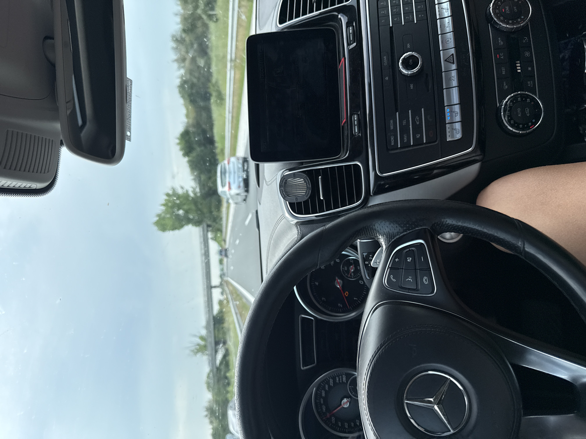 The image depicts the interior of a Mercedes-Benz vehicle, specifically the driver's perspective, on a highway in Gabarnitsa, Bulgaria. The steering wheel, adorned with the Mercedes-Benz logo, occupies the lower left quadrant, displaying multi-function controls on its right spoke. Behind the steering wheel, the instrument cluster shows a speedometer with the needle positioned between 100 and 120 km/h, and a tachometer with an illuminated warning light. To the right, a black infotainment screen is mounted on the dashboard, flanked by an air vent containing a cylindrical "Brooklyn Garage" air freshener. Below the screen, the center console features controls for radio, media, navigation, telephone, and climate control, with the heated driver's seat function visibly active (three blue lights). A driver's bare arm is partially visible in the bottom right corner.
Through the windshield, a multi-lane highway is visible under an overcast sky. The road is bordered by green trees and foliage. An overpass bridge crosses the highway in the mid-distance. Several other vehicles are present on the highway, with a silver hatchback discernible in the lane to the right, further ahead. A rear-view mirror is mounted at the top center of the windshield. The overall environment suggests driving on a rural or intercity highway during daytime.