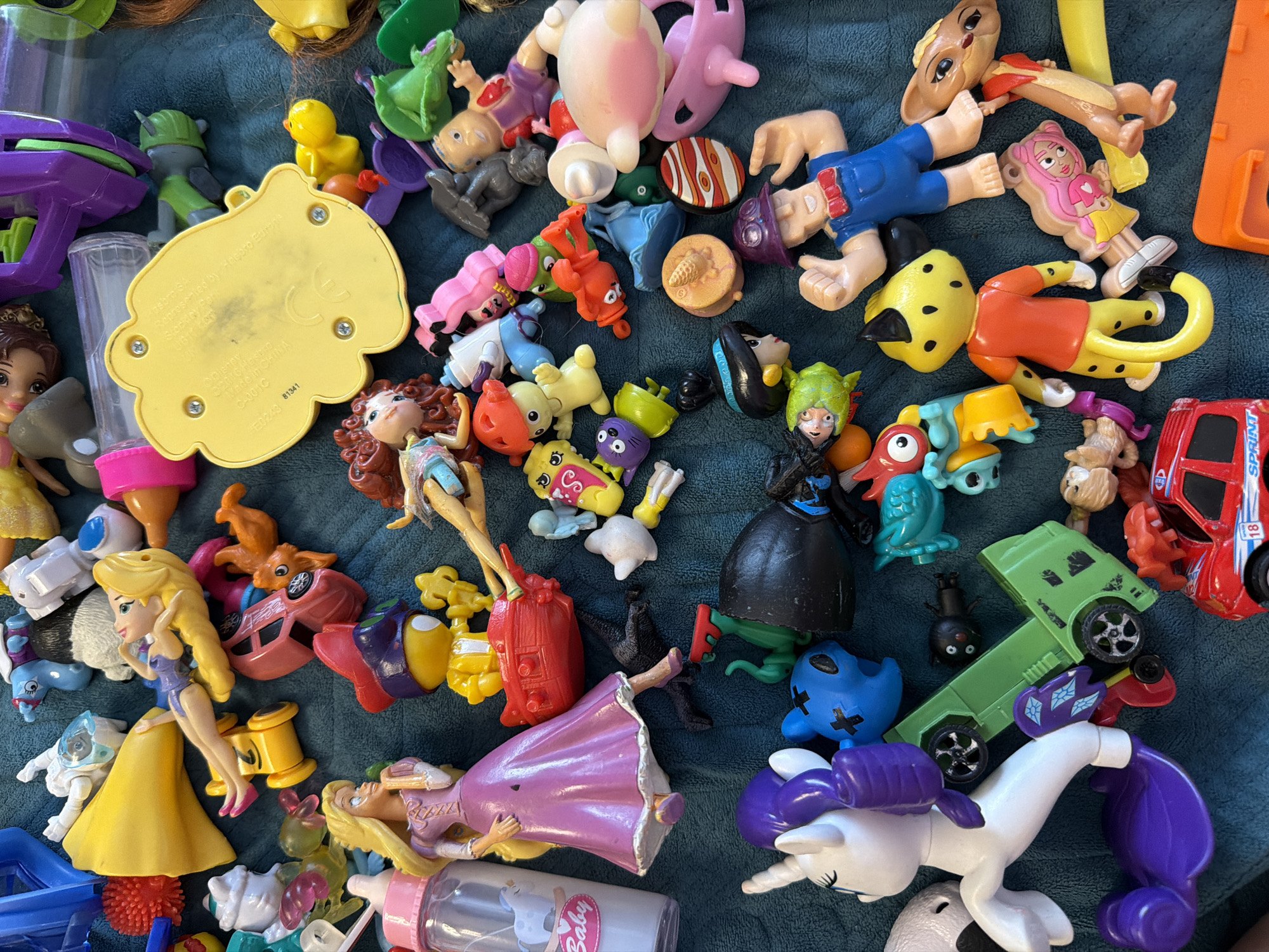 The image shows a collection of various toys scattered on a teal fabric surface. The toys include figurines of Disney princesses (Aurora, Belle), a My Little Pony unicorn, small animal figures, various toy vehicle components, including a red "SPRINT" car, and other miscellaneous plastic toy parts. The toys are arranged haphazardly, overlapping each other.
The city of Sochi, Russia is not relevant to the content of the image.