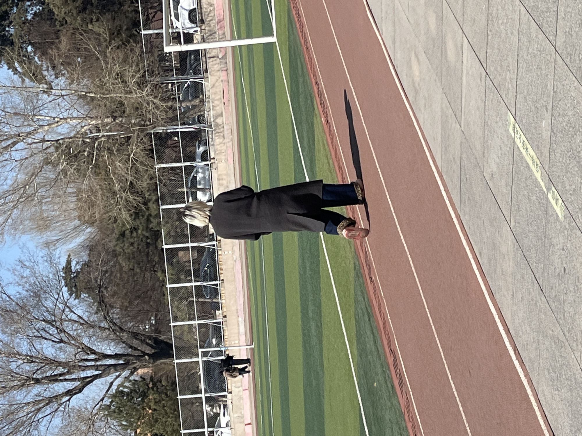 A medium, wide shot depicts a person in a long, dark coat walking on a red track next to a green field in Changchun City, China. The person has light-colored hair. The track is next to a sidewalk with gray tiles. A bare tree and a fence with cars parked behind it are visible in the background. The sky is blue. The scene is well-lit with a long shadow cast by the person.