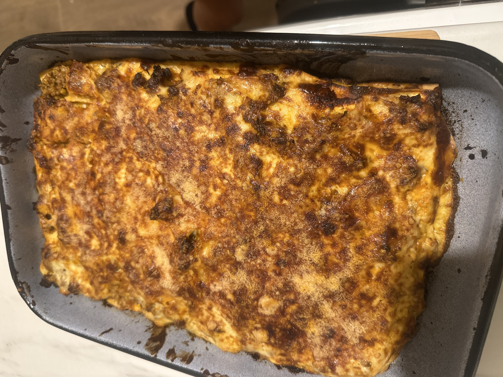 A close-up, overhead shot shows a rectangular baking dish filled with a golden-brown, bubbling lasagna. The cheese on top is melted and slightly browned in spots, with a dusting of what appears to be breadcrumbs or parmesan cheese. The edges of the lasagna show layers of pasta and filling. The baking dish is dark grey and appears to be metal. The setting is indoors, on a light-colored countertop, with a hint of a white appliance or surface on the right side of the frame. No people are visible in the image. The location is not specified within Svilengrad, Bulgaria.