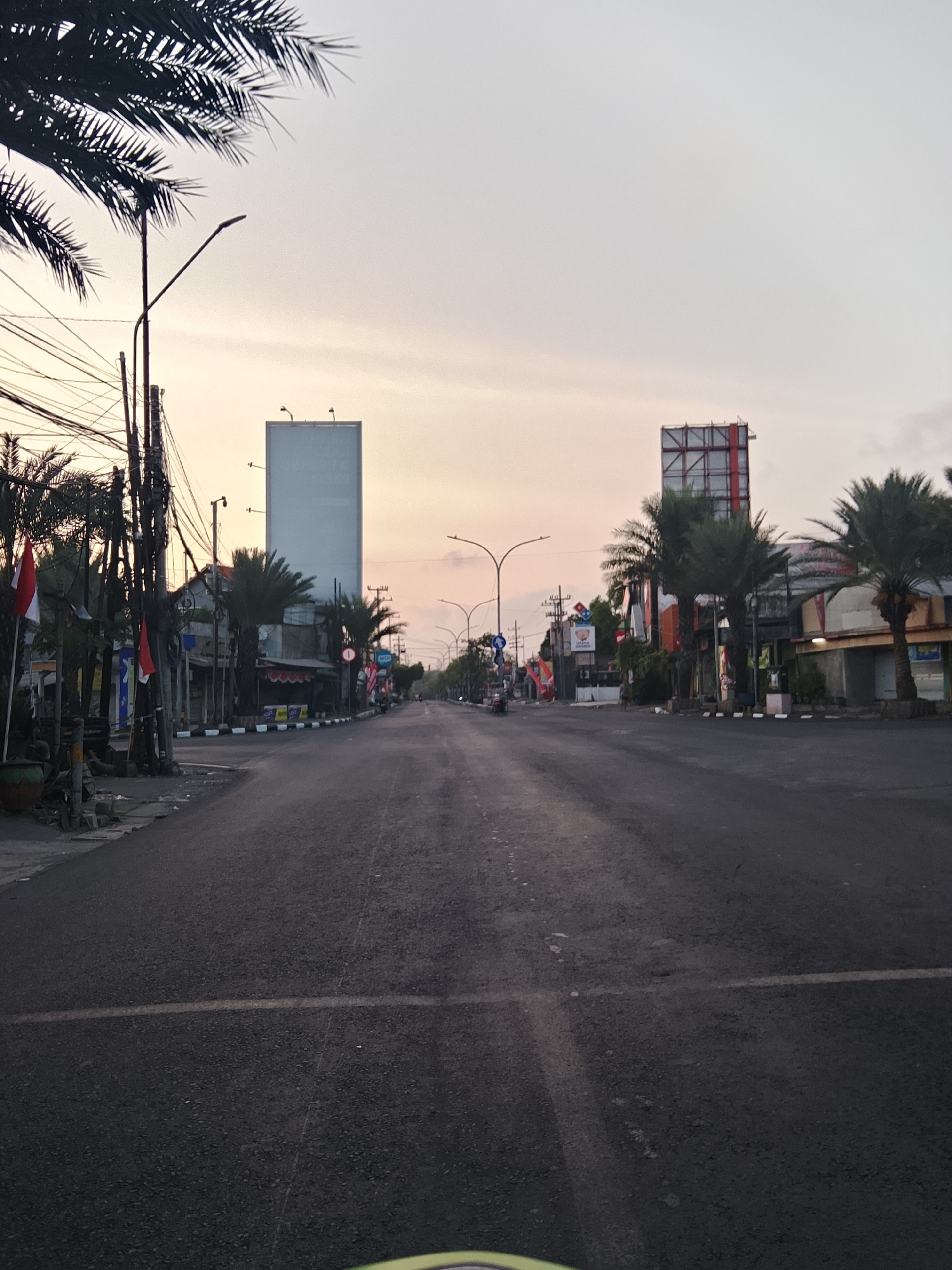 The image depicts an empty street in Sukorame, Indonesia, at either dawn or dusk, with soft, warm lighting casting long shadows. The road stretches towards the horizon, flanked by buildings and palm trees. On the left side of the road, Indonesian flags are visible, along with several utility poles and a large billboard. The right side of the road also features buildings, palm trees, and another billboard. The sky is a pale, diffused orange and white, indicating the time of day. No people are present in the immediate foreground, but distant figures are faintly visible further down the street. The overall impression is one of tranquility and quietude.