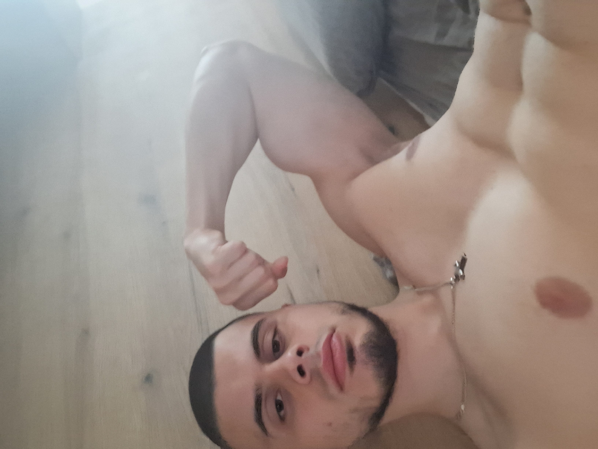 A shirtless, light-skinned male lies on a wooden floor. He has a buzz haircut and a short, dark beard. He is wearing a silver chain necklace. His right arm is flexed upwards. The location is not discernible, so providing any details about Burgas, Bulgaria is impossible.