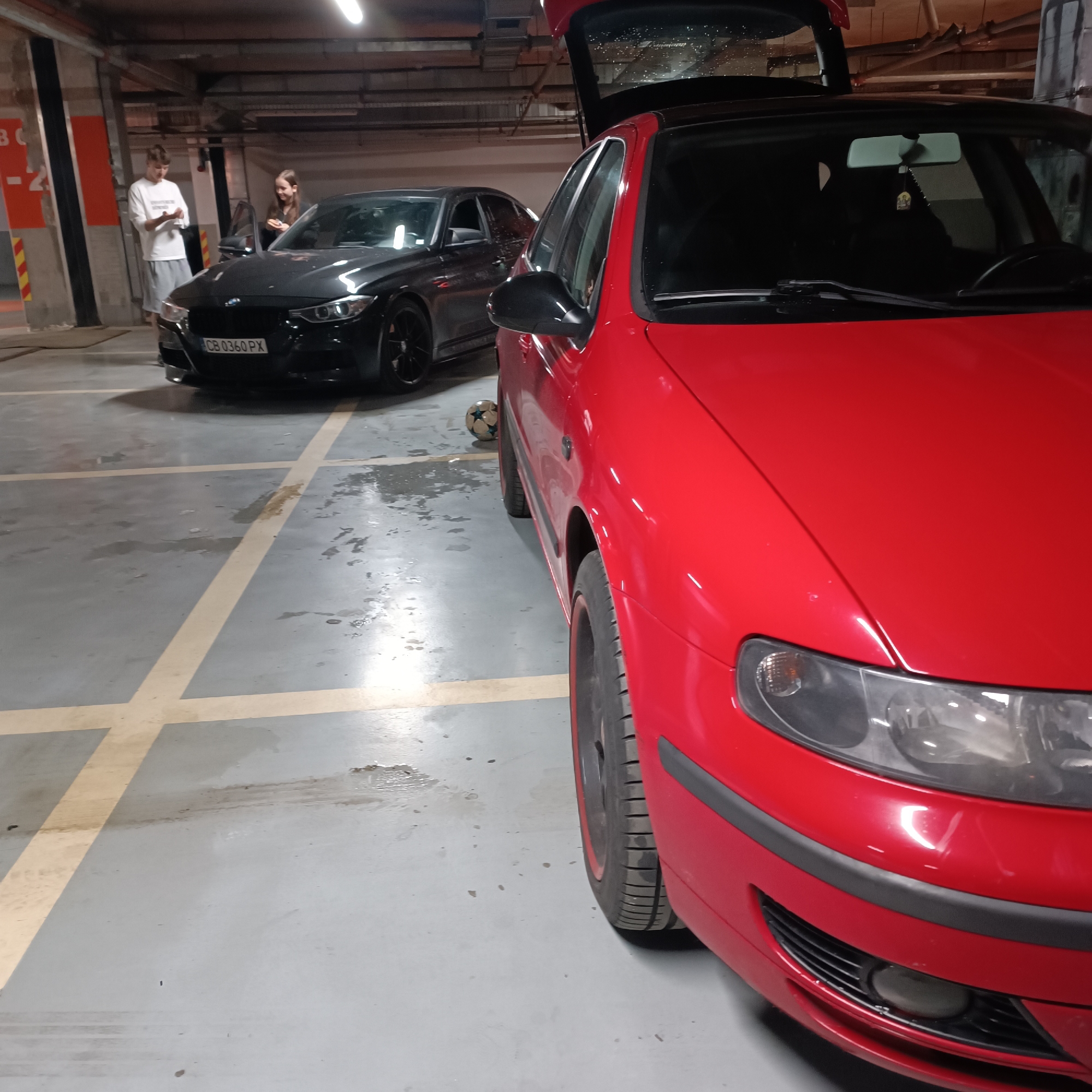 The image depicts an indoor parking garage with artificial lighting. Two cars and two young individuals are the main subjects.
In the foreground to the right, a red car is partially visible, facing the viewer's left. Its front right side, including the headlight, bumper, and a tire with a black rim and red accent, is in focus. The car's trunk is open, revealing the interior.
Further back and to the left, a black car, appearing to be a BMW sedan, is parked with its driver's side door open. A male figure, wearing a white t-shirt and grey shorts, stands near the front of the black car, looking down at something in his hands. Behind the black car, a female figure, dressed in dark clothing, is visible, looking towards the male and also holding something.
Between the two cars, a black and white soccer ball rests on the concrete floor. The floor itself is concrete with yellow painted lines marking parking spaces, and shows some wet patches and dirt marks. A large concrete pillar, partially painted red or orange, with the text "B -2" is visible on the far left. The ceiling of the garage shows exposed ductwork and lighting fixtures.
The license plate "CB 0360 PX" is clearly visible on the front of the black car, indicating a location in Sofia, Bulgaria. The overall atmosphere is casual, suggesting people are hanging out or preparing to leave from the parking garage.