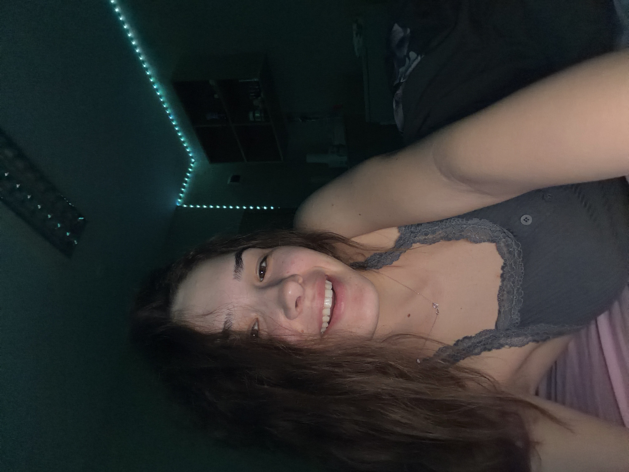 The image displays a young woman, likely in her late teens or early twenties, captured in a close-up, horizontal selfie. She is smiling broadly with a cheerful expression, revealing her teeth, and her light brown eyes are open and looking towards the camera. Her long, dark brown hair cascades around her face and extends across the bottom of the frame. She wears a dark grey or charcoal-colored camisole with a delicate lace trim along the neckline, and a thin silver necklace with a small pendant is visible on her neck.
The setting appears to be an indoor room, likely a bedroom, characterized by low light conditions suggesting it is nighttime or late evening. The primary illumination comes from teal or blue LED strip lights that line the edges of the ceiling in the background. Faint details of shelves or wall-mounted storage units are discernible in the deep shadows behind her. A glimpse of pink or purple fabric, possibly a bedsheet or blanket, is visible on the far right. The woman's pose and genuine smile convey a relaxed, happy, and informal mood. No visible text is present in the image. The location context provided for this scene is Sofia, Bulgaria.