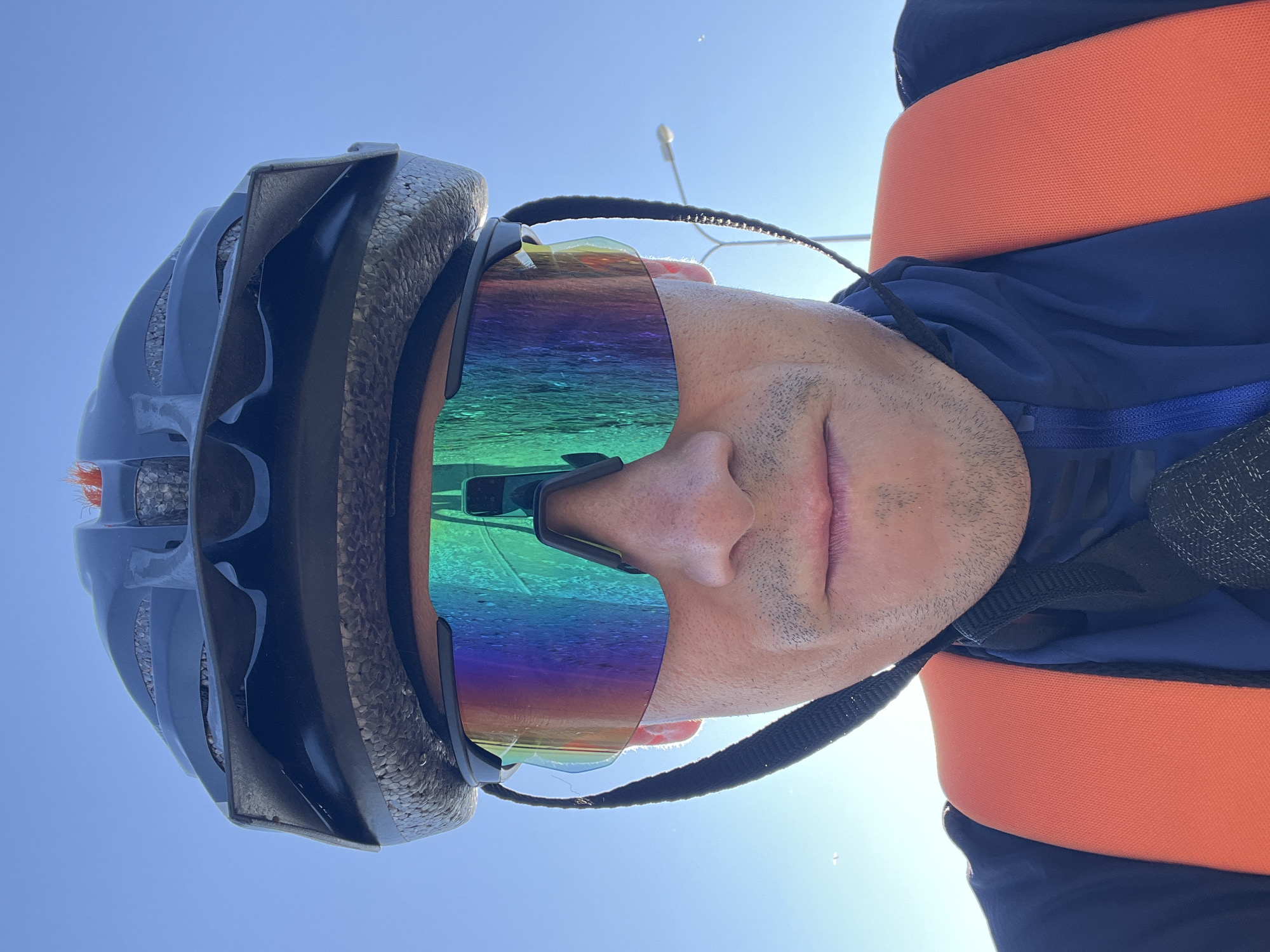 A close-up, forward-facing, low-angle, selfie-style photo shows a person wearing a black bicycle helmet and large, iridescent, mirrored cycling sunglasses. The sunglasses reflect the sky and a portion of the person's face. The person is wearing an orange vest over a dark blue jacket. The background is a clear blue sky with a faint hint of a lamppost on the right side. The image is captured outdoors during the daytime, likely in a public area within Chaikovsky, Russia.