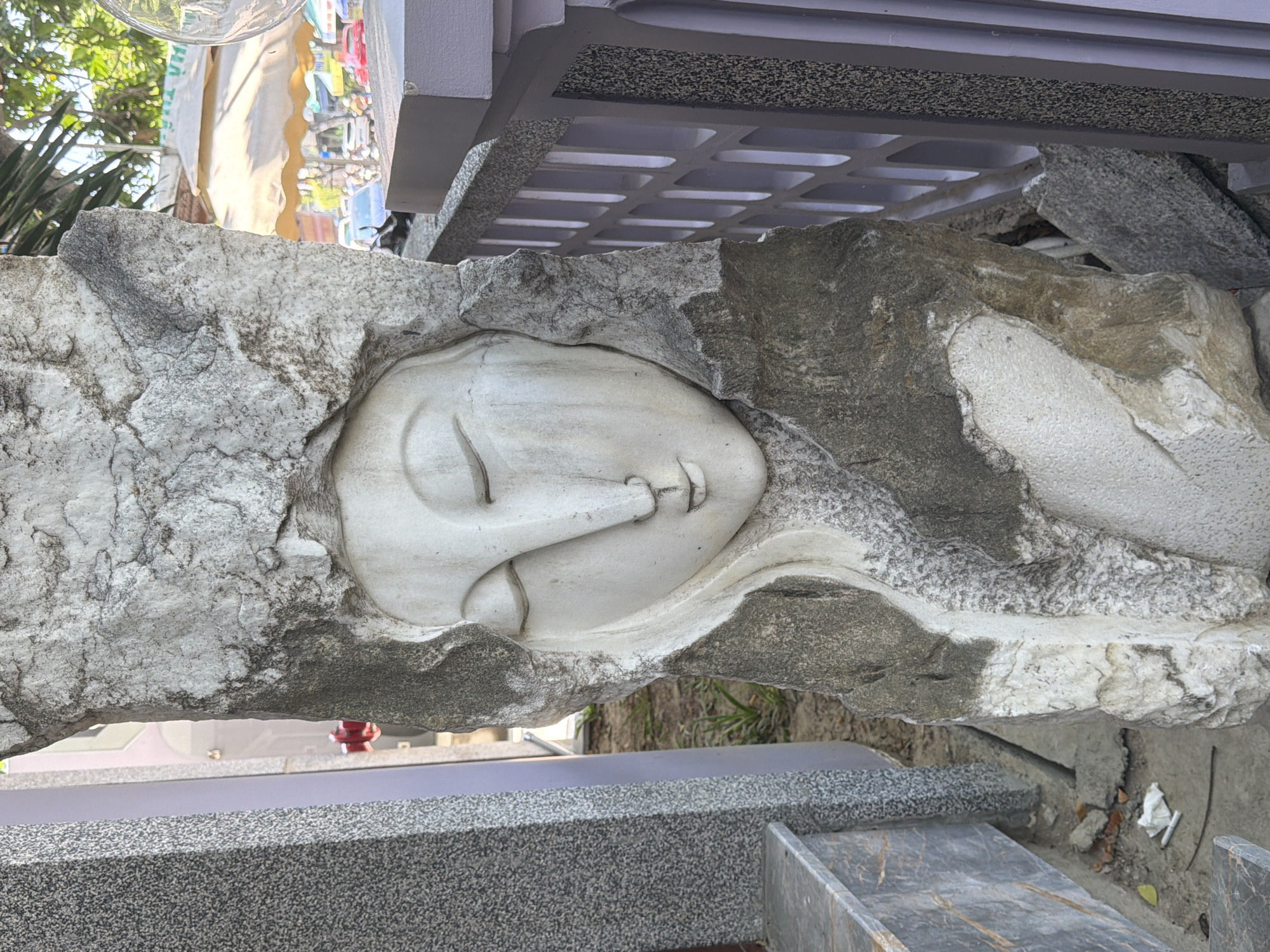A close-up, eye-level, full shot captures a stone sculpture of a serene face, with closed eyes and a slight smile, carved into a rough, grey, and white stone pillar. The sculpture is positioned outdoors, to the right of a speckled grey gravestone-like structure and to the left of a pale lavender fence with a patterned opening. In the background, a busy street is visible with cars, awnings, and greenery, suggesting an urban setting within Nam Nha Trang Ward, Vietnam. The lighting is bright, casting soft shadows and highlighting the texture of the stone.
