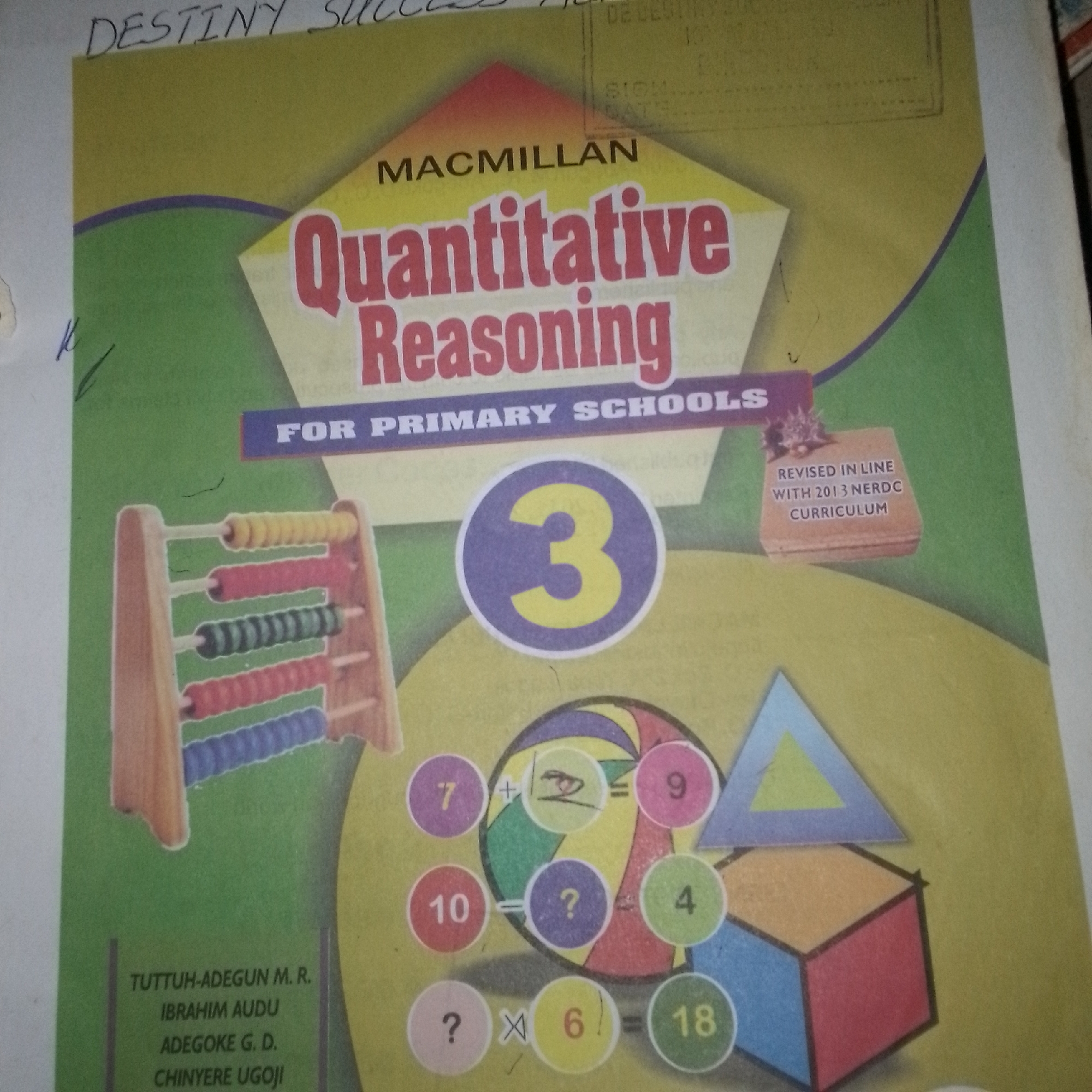 The image is a close-up shot of the cover of a book titled "Quantitative Reasoning For Primary Schools" by Macmillan. The book is yellow with a large purple circle containing a yellow number 3 prominently displayed. There are various mathematical elements depicted, including an abacus, geometric shapes, and simple equations with numbers and question marks. The book also mentions "REVISED IN LINE WITH 2013 NERDC CURRICULUM". The bottom left corner of the cover lists authors: "TUTTUH-ADEGUN M. R., IBRAHIM AUDU, ADEGOKE G. D., CHINYERE UGGOJI". The background is a plain yellow color, suggesting this is a book cover rather than a photograph of a scene. There is no indication or visual evidence within the image that places it in Jalingo, Nigeria, or any other specific city.