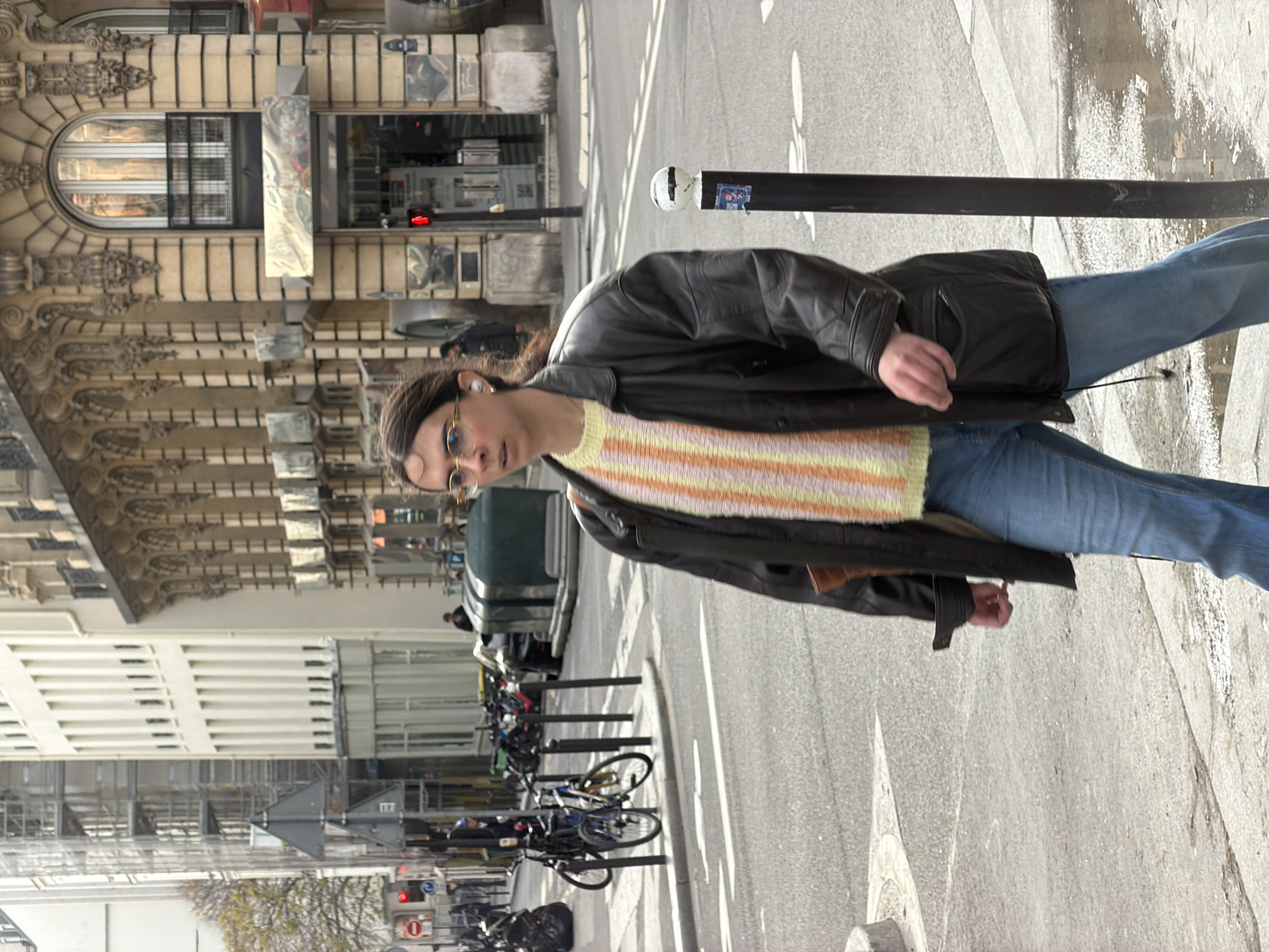 A person with long brown hair, wearing eyeglasses, a striped pastel sweater, and a black leather jacket, is standing on a sidewalk next to a street. The person is wearing blue jeans. The sidewalk is made of concrete and has a narrow, dark post near the edge.
On the other side of the street, a row of bicycles are parked. Beyond the bicycles is a pedestrian crosswalk. In the background, a building with ornate architectural details can be seen. A large window with multiple panes is visible.
The scene is set in Paris, France. The building architecture is typical of the city. The overall lighting suggests a daytime scene with diffused sunlight.
