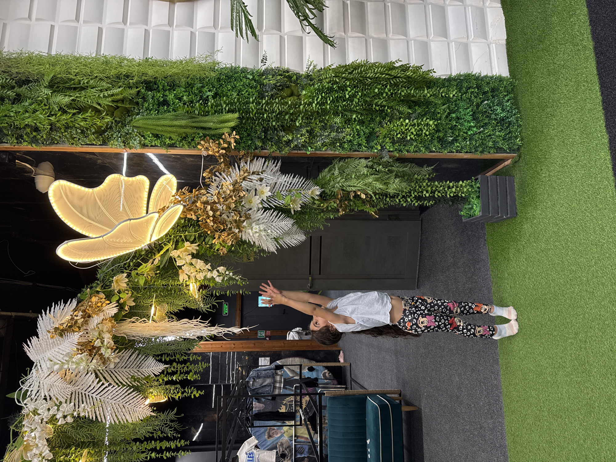 A young girl with dark, curly hair, wearing a white t-shirt and patterned leggings, stands on a patch of artificial grass, reaching her hands upwards. She is looking at a large, illuminated butterfly decoration. Behind her, there is a dark doorway and clothing racks with garments. To her right, a vertical garden wall of lush green plants is mounted next to a white, textured wall. The scene appears to be an indoor space, possibly a shop or an event venue, with the artificial turf suggesting a playful or decorative element. The location is not identifiable as Sochi, Russia.