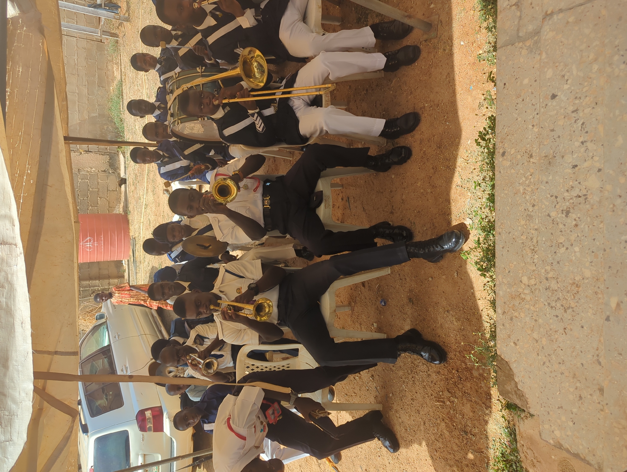 A group of young people, primarily male, are seated outdoors playing brass instruments. They appear to be part of a band, possibly a school or community ensemble. The individuals are wearing uniforms, some with white shirts and others with black and white jackets or coats. They are seated on white plastic chairs.
In the background, there is a parked white vehicle and a reddish-orange barrel or container. The setting appears to be a semi-outdoor area, possibly under a makeshift tent or awning. The ground is dirt, and there's a concrete structure to the right.
Based on the request, the location is Sabon Tasha, Nigeria.