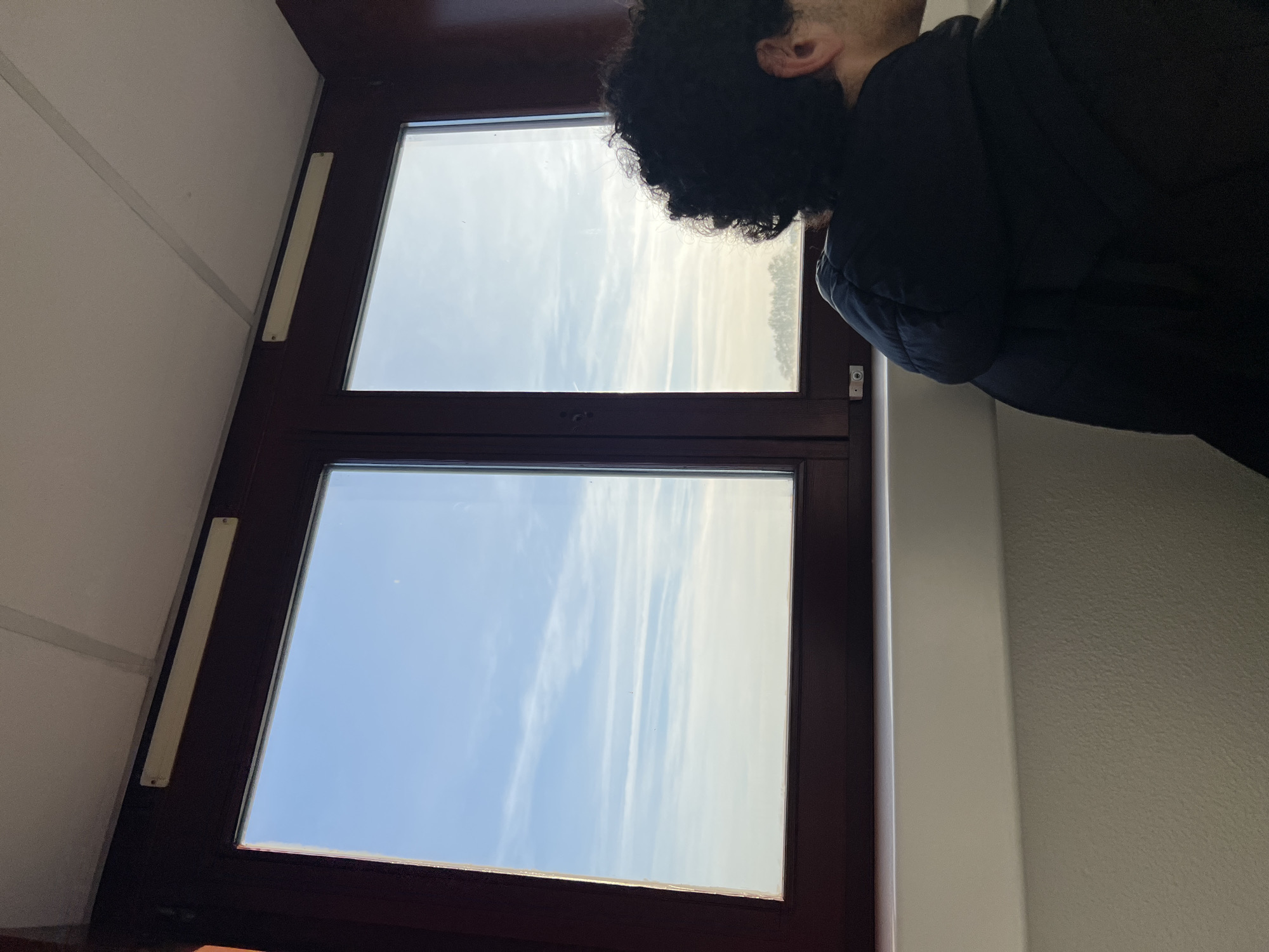 A person with curly hair and a black jacket is looking out of a brown-framed window. The window is composed of two panes. The sky is visible through the window, with some wispy clouds. The location is Schiltigheim, France.