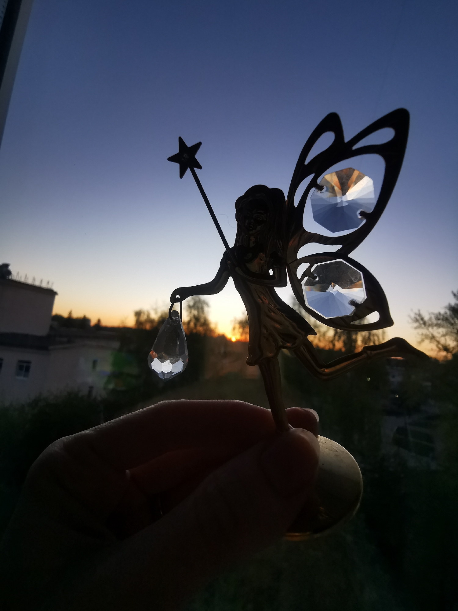 A hand holds a fairy figurine in silhouette against a sunset sky. The fairy figure has butterfly-like wings made with cut crystals, a star-topped wand, and holds a crystal pendant. In the background, buildings and trees can be seen. Based on metadata, the location is Chaikovsky, Russia. The photo was taken indoors looking out a window.