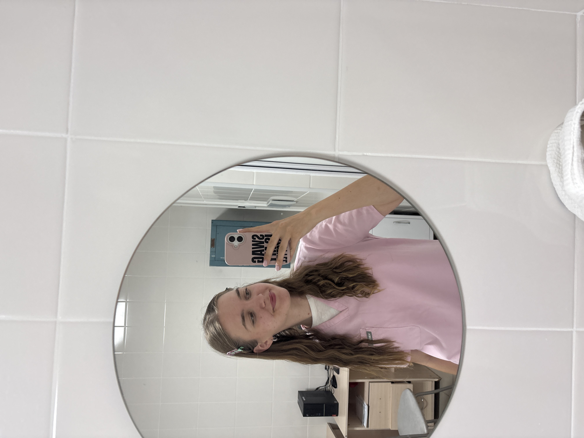 In a bathroom with white tiled walls, a young woman with wavy brown hair and two colorful hair clips takes a selfie in a round mirror. She is wearing a pink scrub top over a white undershirt and has bright pink fingernails. Her phone, with a pink case displaying the word "JAWS" in black letters, is visible in her reflection. To her left, a black computer tower sits on a wooden desk, and a stool with a grey seat is partially visible. In the background of the reflection, a blue cabinet is mounted on the wall. The scene appears to be a private or institutional bathroom, possibly in a medical or educational facility, as suggested by the scrubs and the utilitarian nature of the room. The overall impression is of a casual, personal moment captured in an everyday setting. The location cannot be determined from the image.